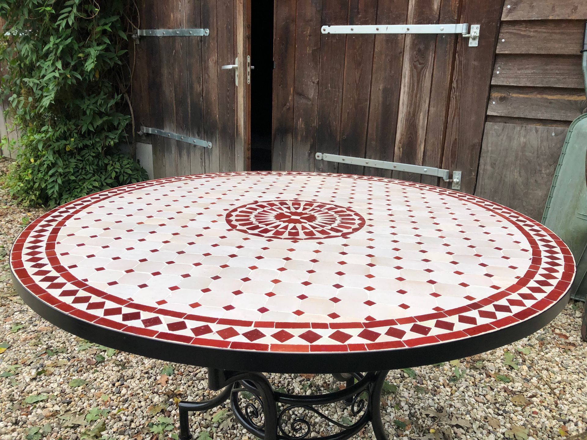 Ronde mozaïektafel met rode en witte tegels, buiten geplaatst bij een verweerd houten gebouw.