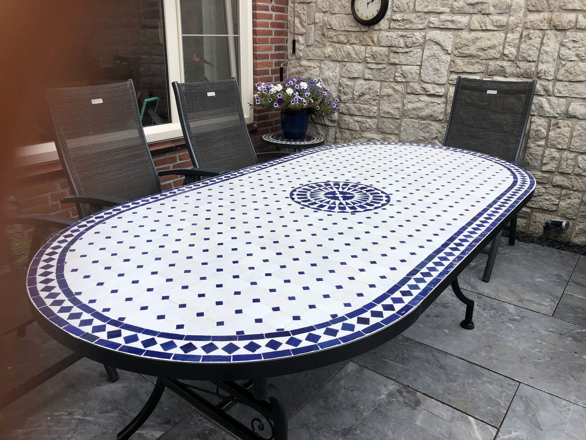 Ovale Marokkaanse terrastafel met een blad van blauw-witte mozaïektegels, omringd door stoelen, op een stenen terras.