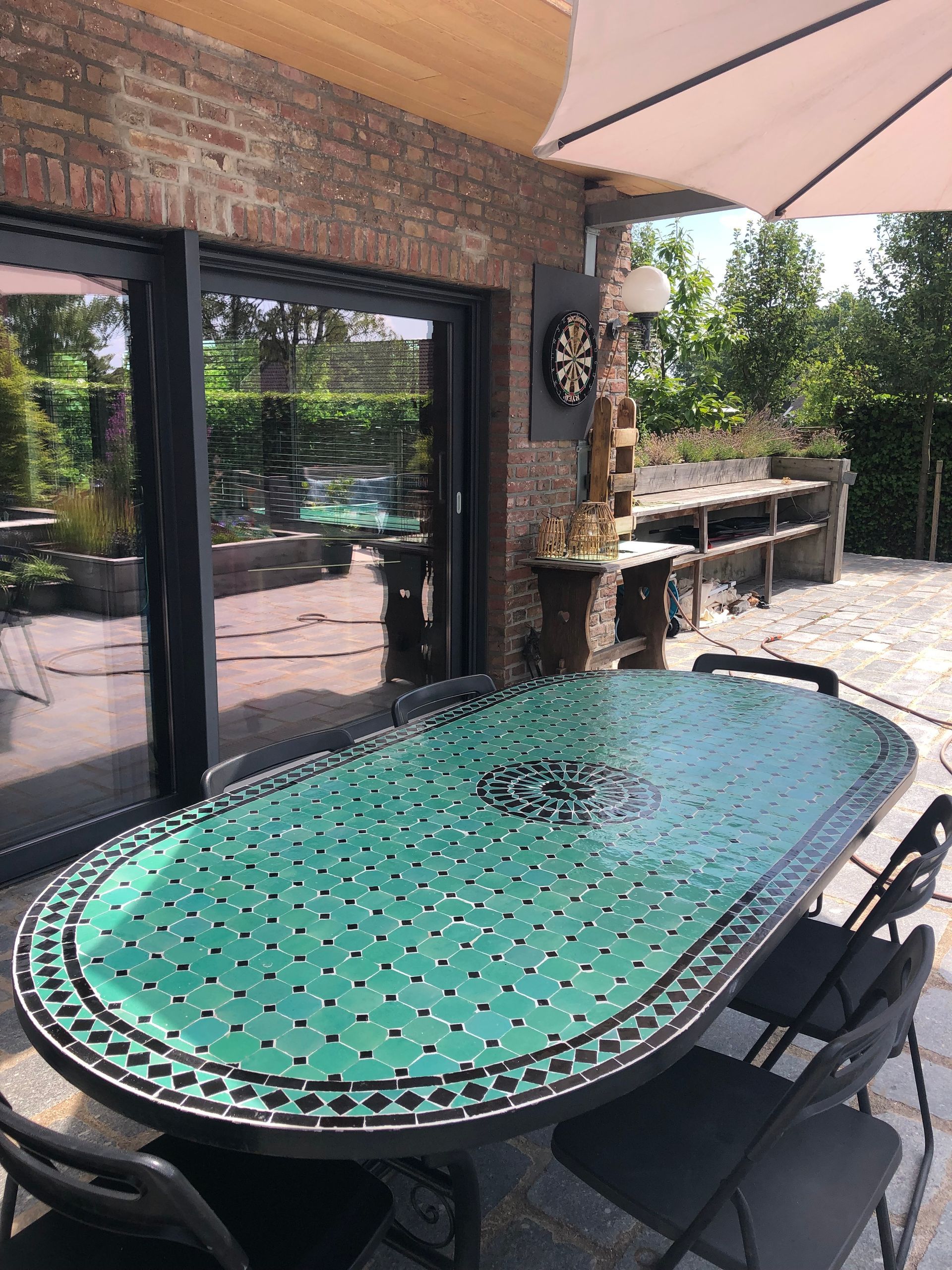 Eettafel voor buiten met een groen mozaïekblad, omringd door stoelen, naast een bakstenen muur en tuin.