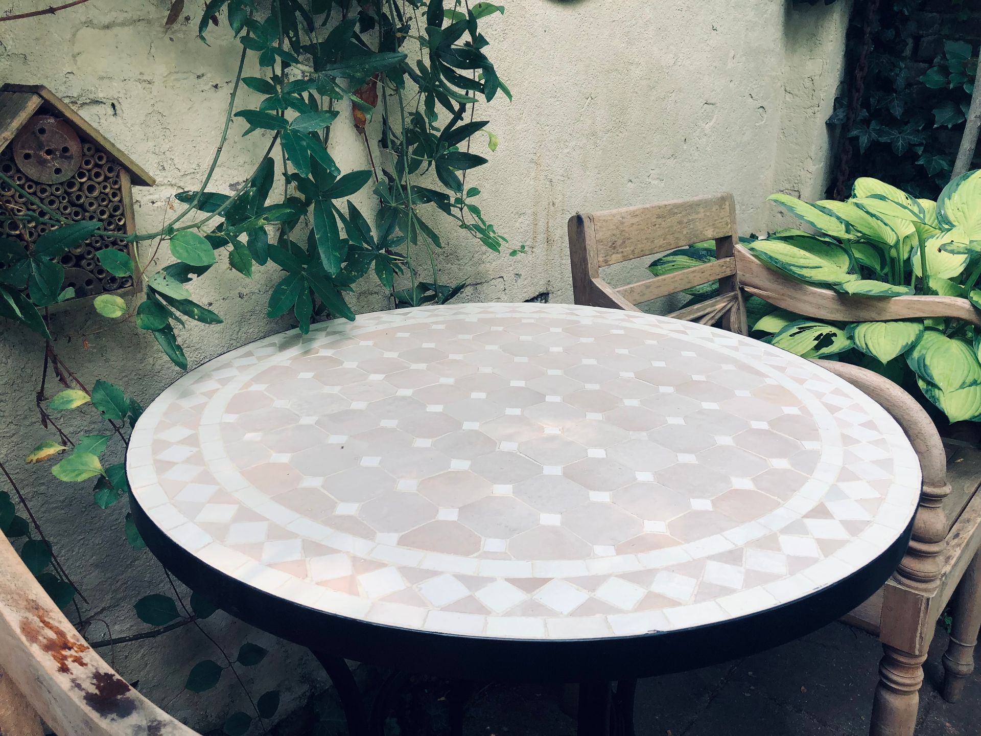 Ronde handgemaakte Marokkaanse  tafel met mozaïektegels en twee houten stoelen op een terras met planten en een bijenhotel.