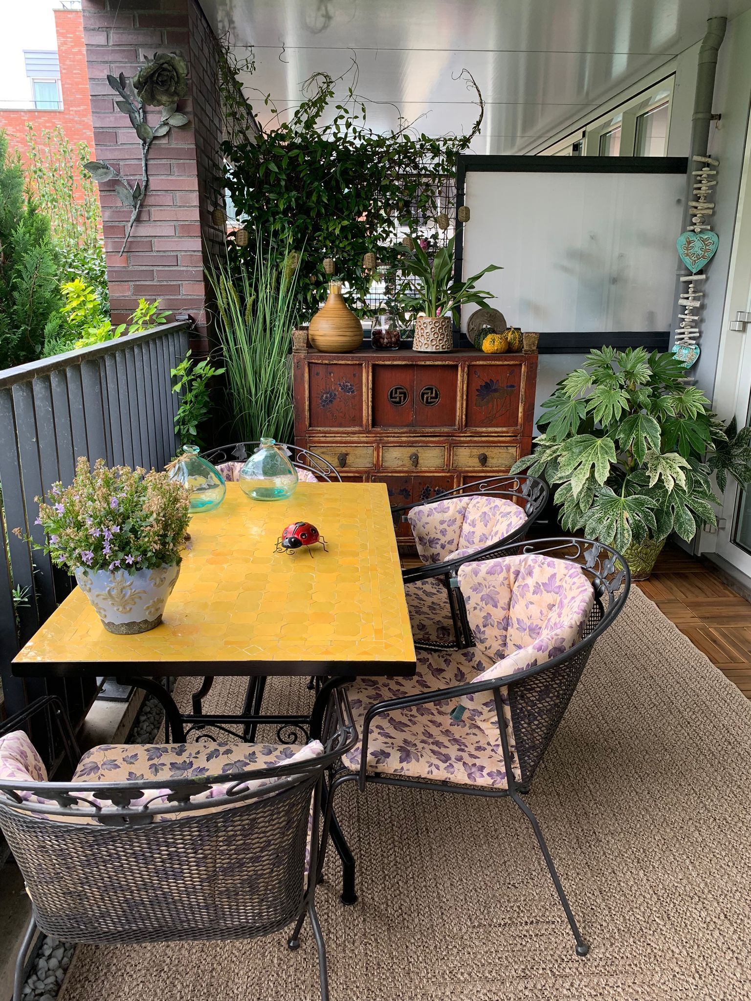 Balkon met tafel, stoelen, planten en een kast. Gele tafel, rieten stoelen en diverse planten.