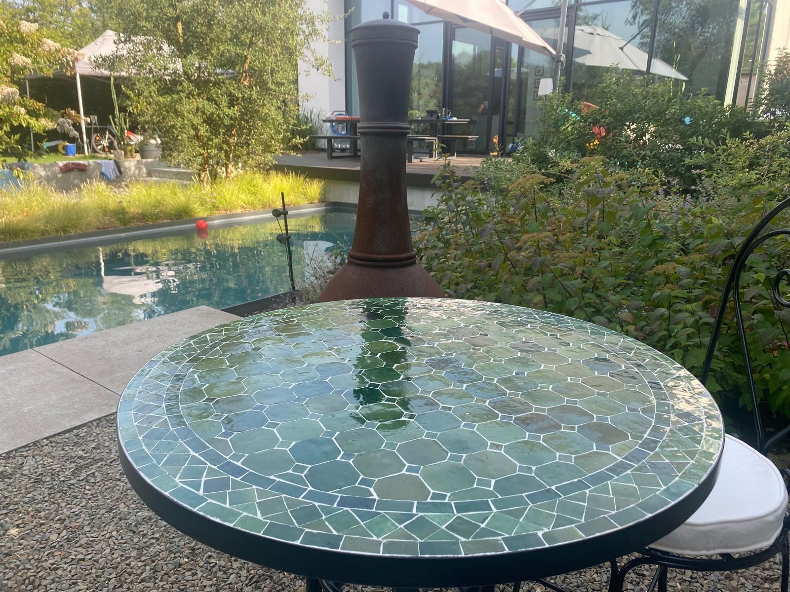 Ronde mozaïektafel uit Marokko met een vijver en groen op de achtergrond. Een vuurplaats in de verte.