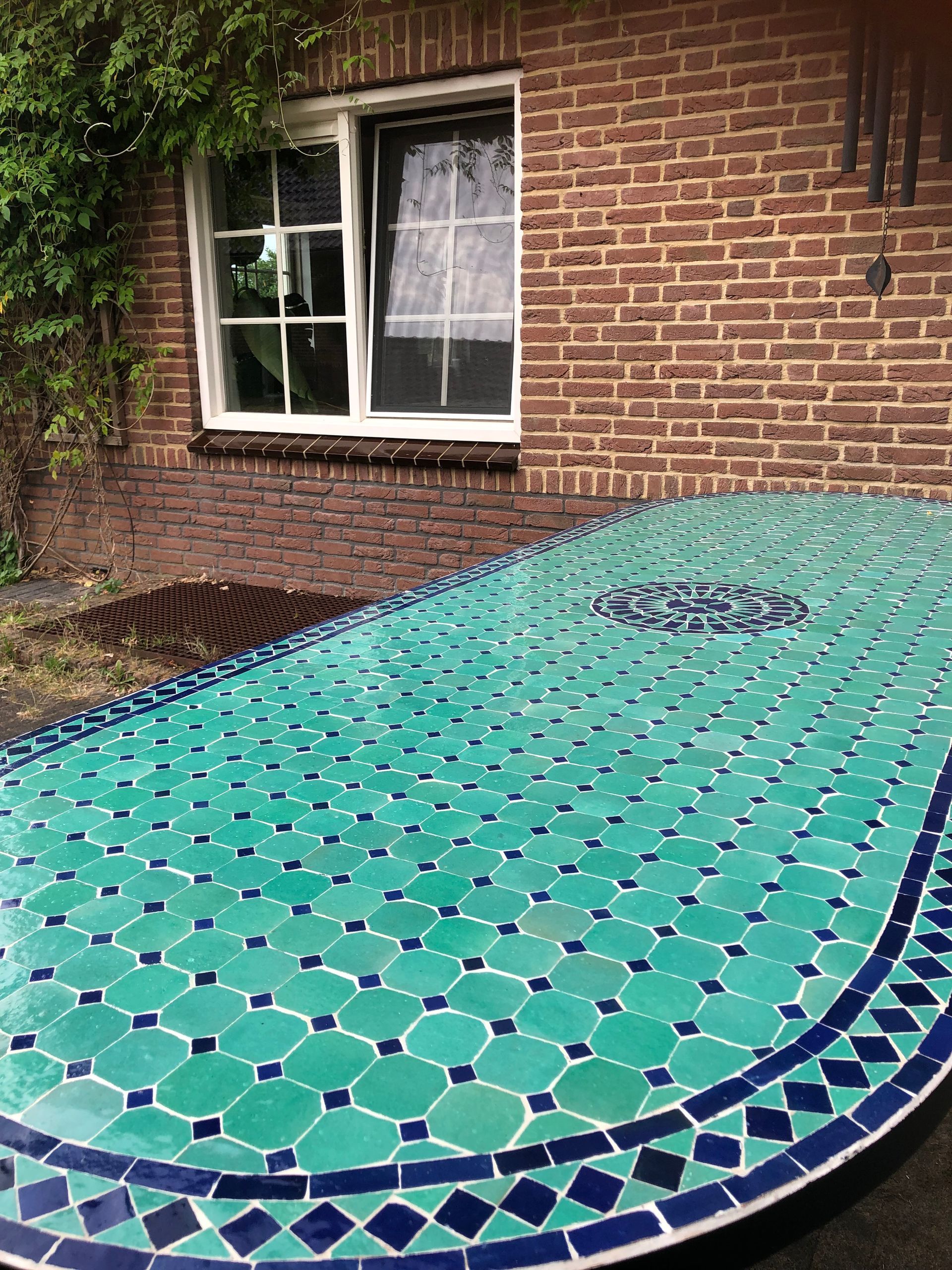 Een turquoise Marokkaanse  tegeltafel staat buiten voor een bakstenen muur en een raam met een wit kozijn.