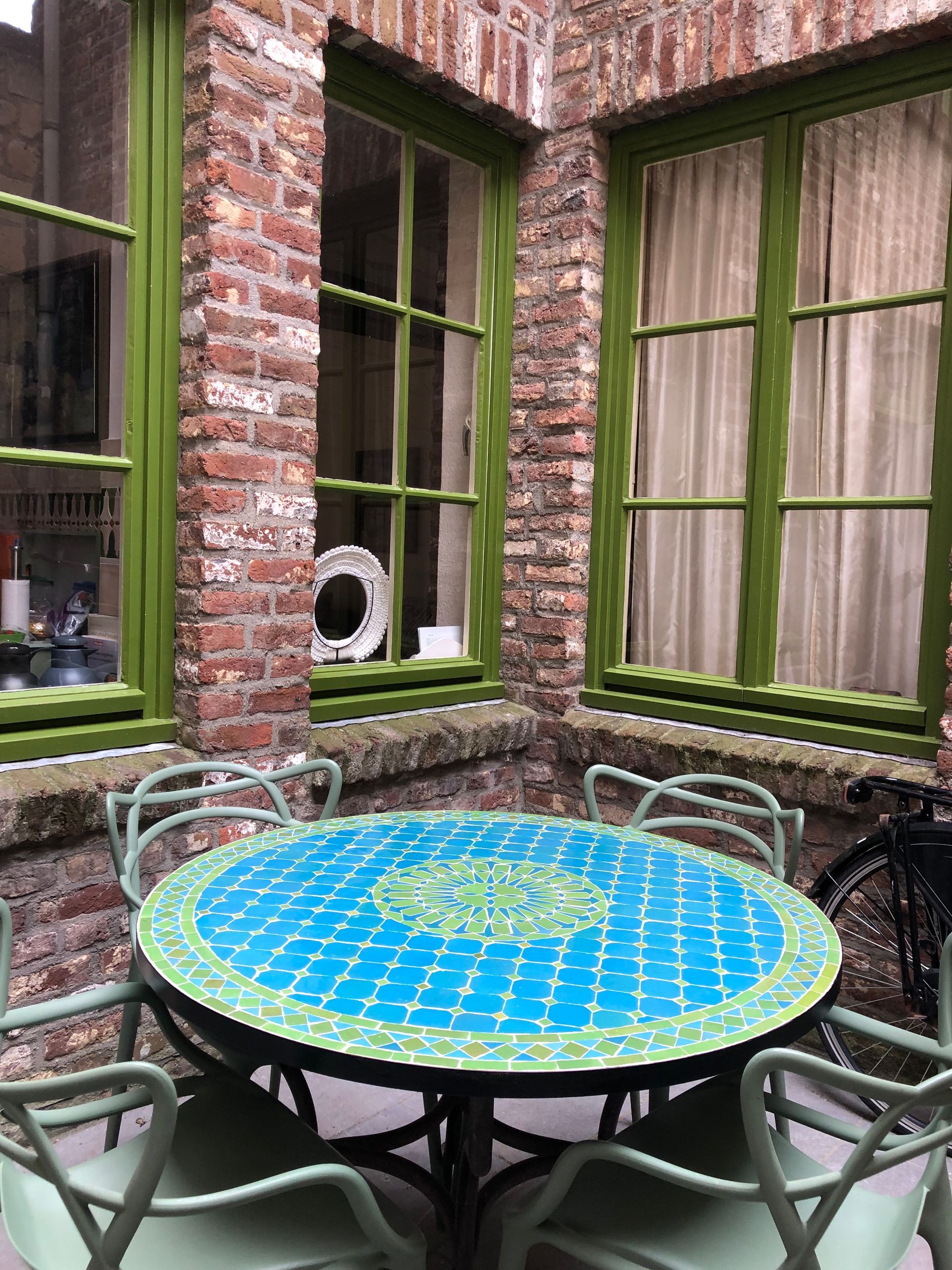 Binnenplaats met een turquoise tafel en groene stoelen, omlijst door groen omlijste ramen en bakstenen muren.