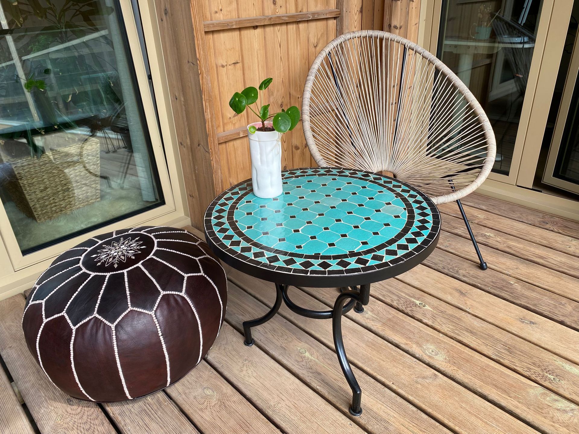 Ronde handgemaakte Marokkaanse tafel met turquoise mozaïekblad, geweven stoel en leren poef op een houten terras.