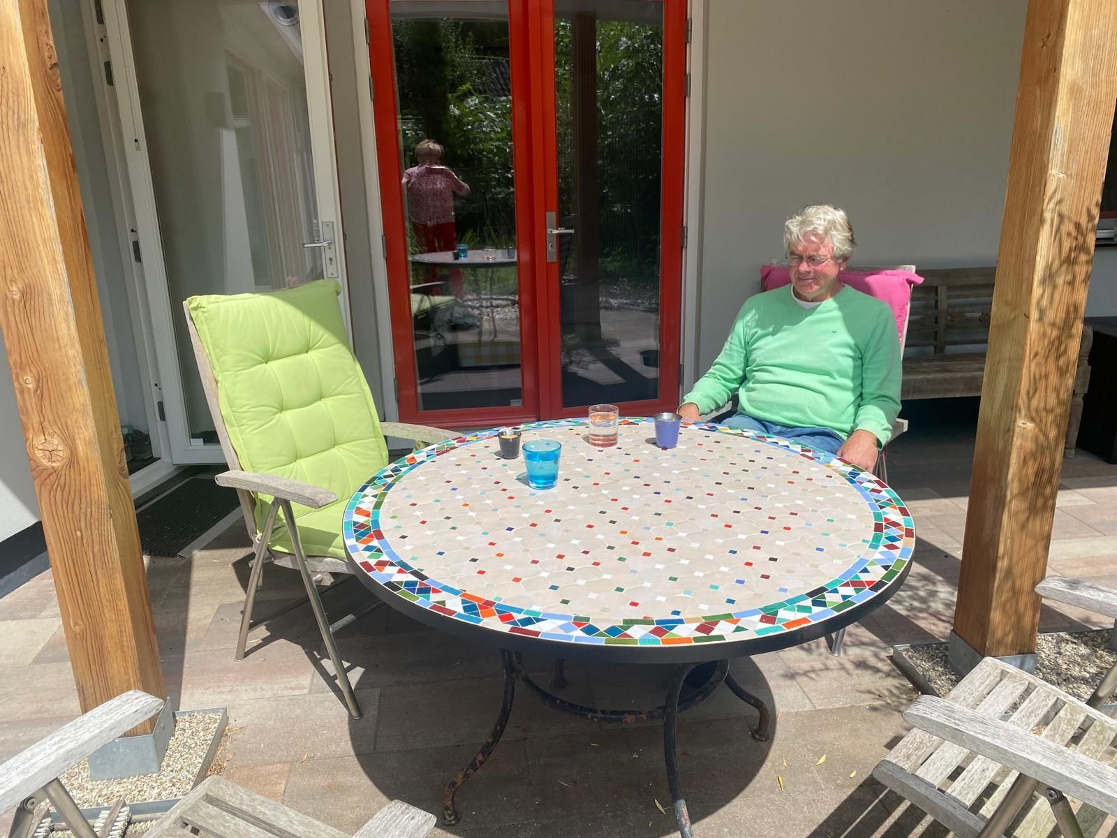 Een man zit buiten aan een prachtige Marokkaanse mozaïektafel. Rode deuren en een groene stoel zijn zichtbaar.