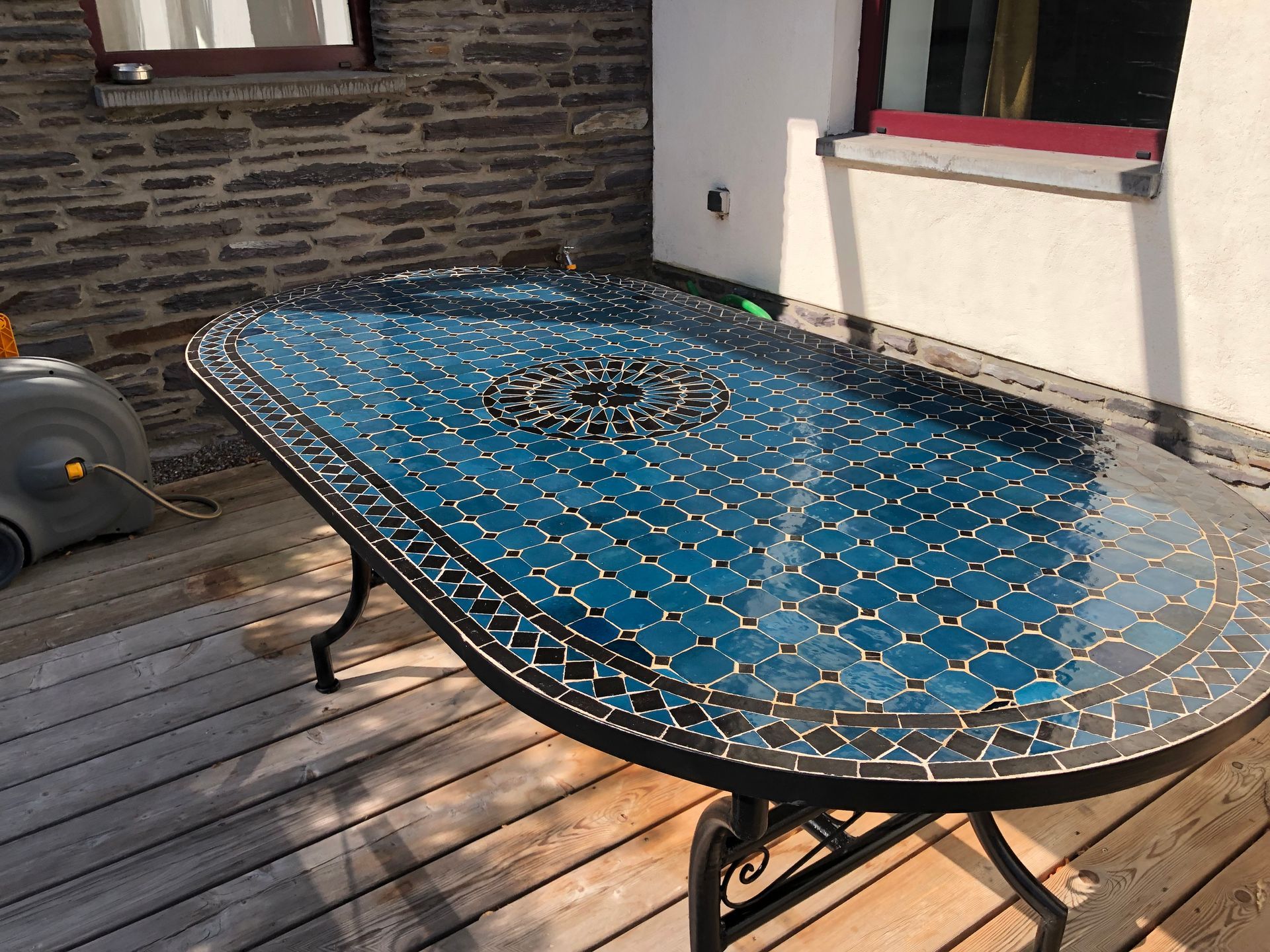 Marokkaanse Tuintafel met mozaïekblad op een houten terras. Blauwe mozaïektegels, zwart frame. Zonlicht.