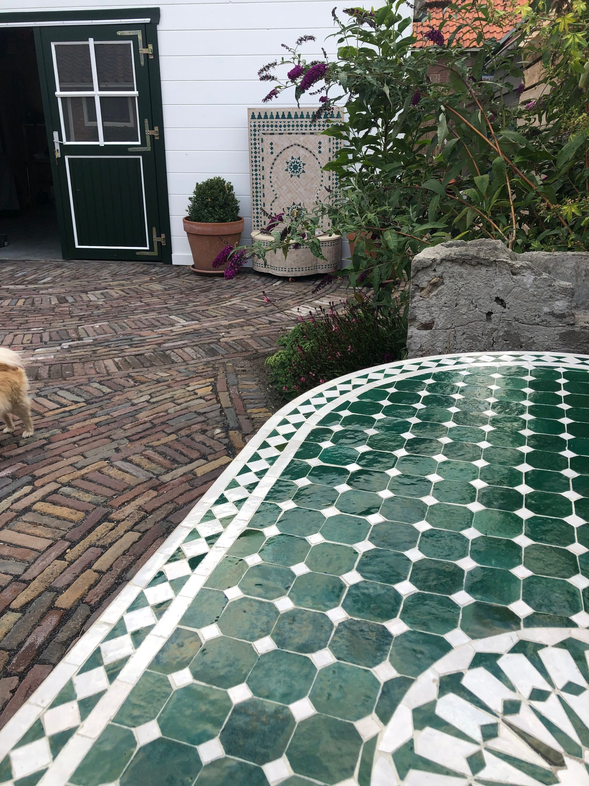 Groene Marokkaanse mozaïektafel op een bakstenen binnenplaats; groene deur en potplant op de achtergrond.
