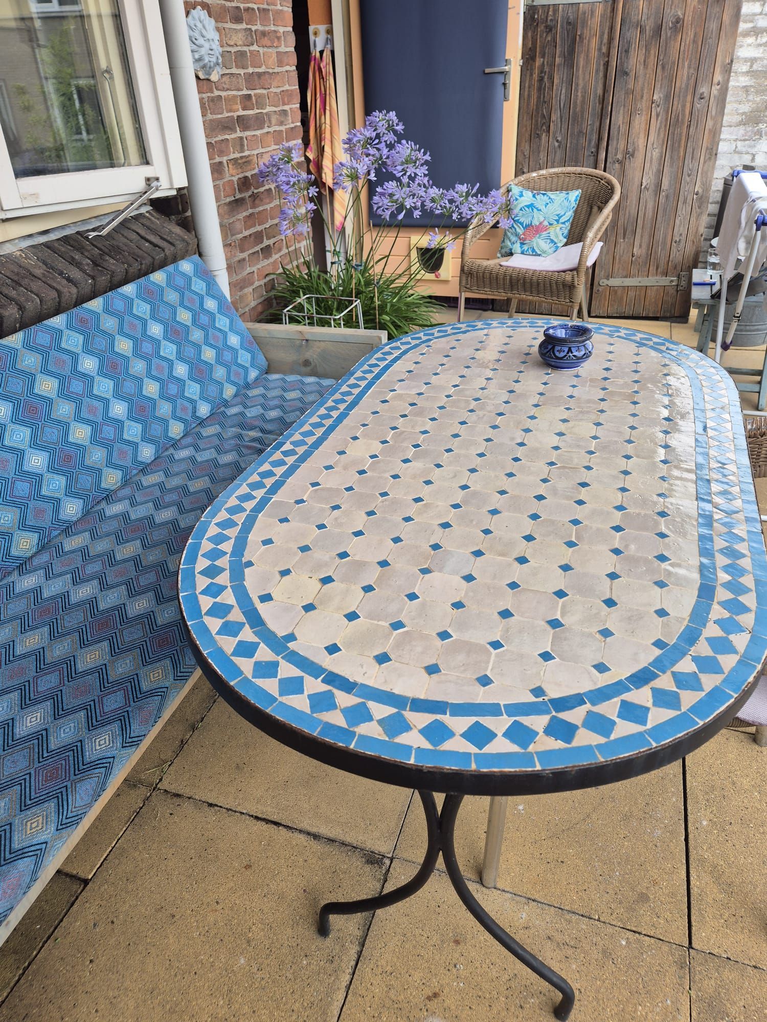 Terras buiten met een Marokkaanse mozaïektafel, blauwe gestoffeerde bank en rieten stoel.