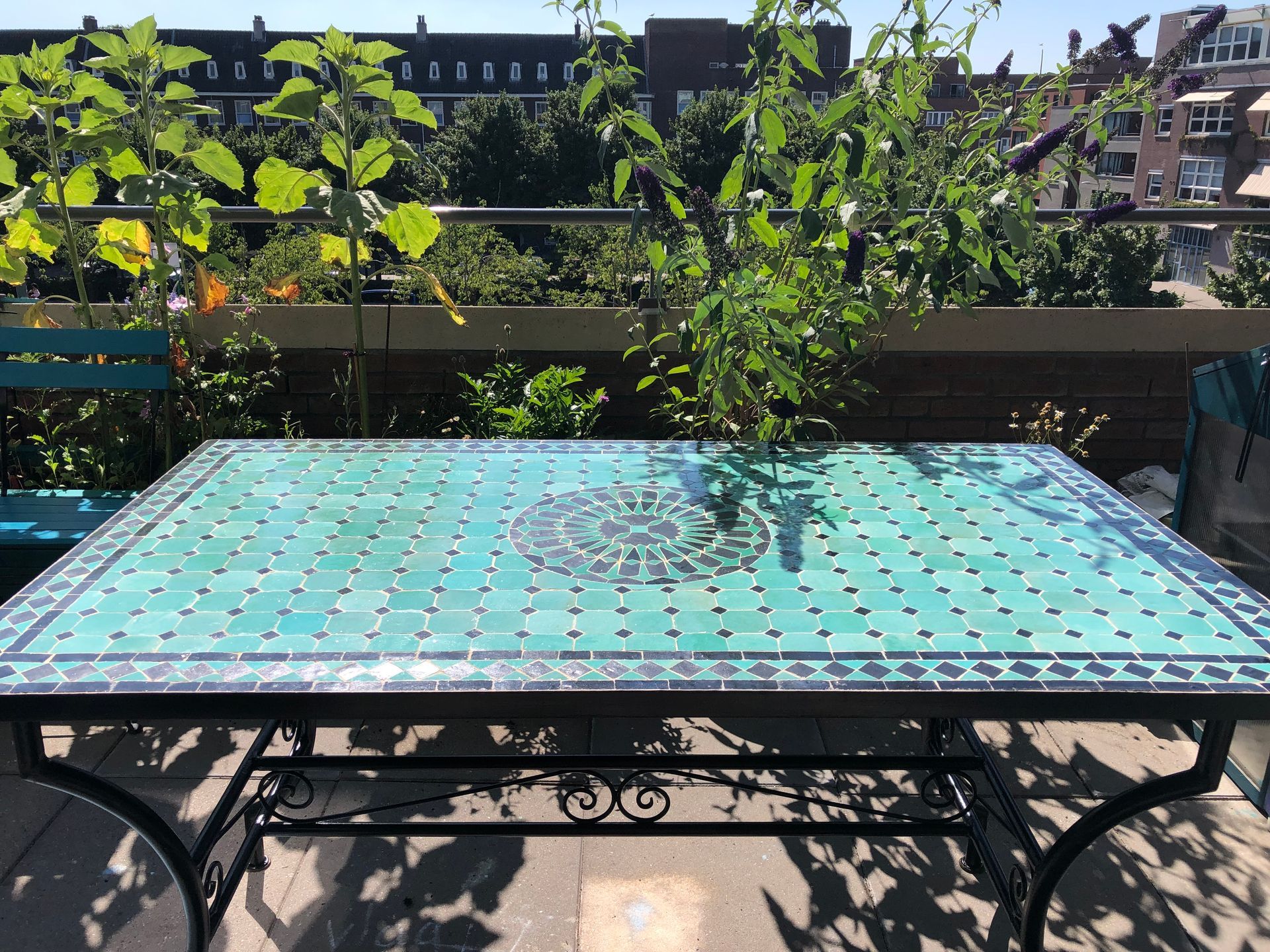 Een turquoise tafel met mozaïektegels op een terras, met groen en gebouwen op de achtergrond.