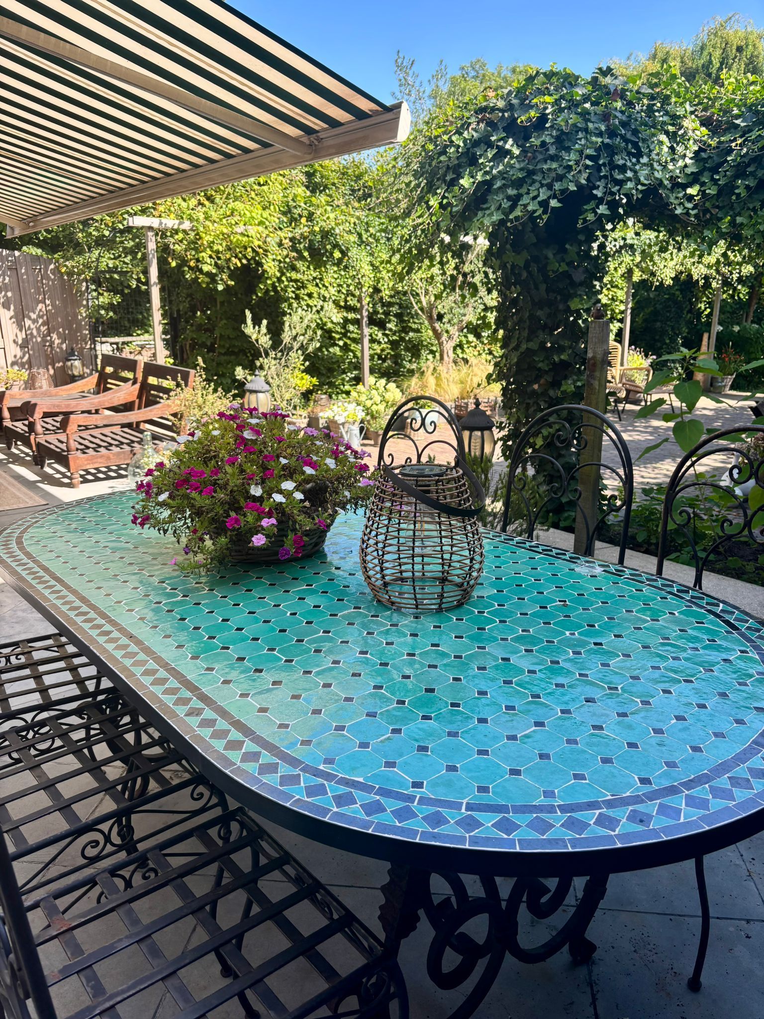 Een eettafel gedekt voor buiten, met een tafelblad van mozaïektegels en metalen stoelen, onder een schaduwrijke pergola.