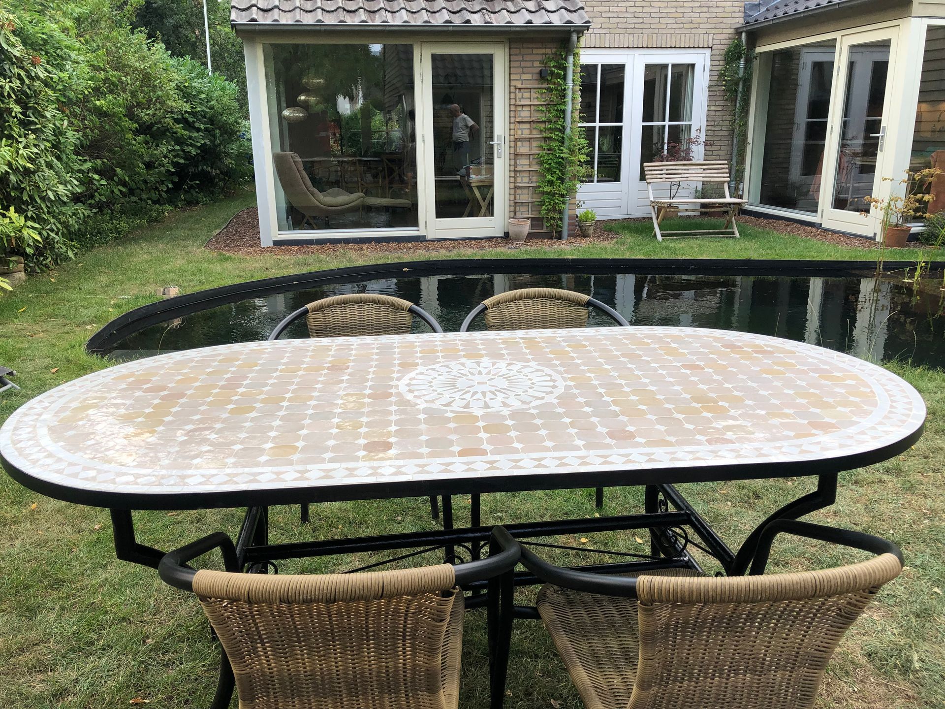 Eetset voor buiten met ovale Marokkaanse mozaïektafel en vier stoelen met gevlochten rugleuning op een grasveld.