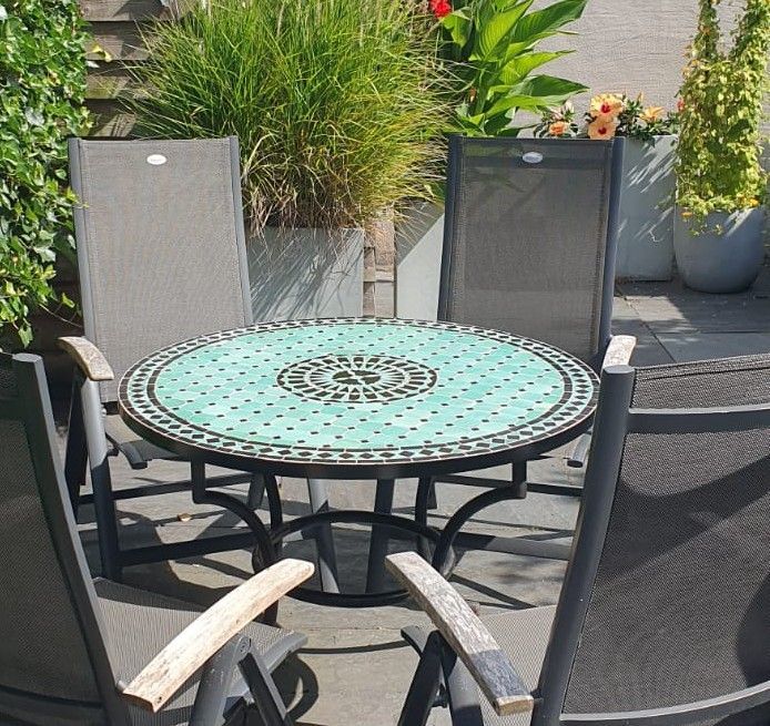 Ronde tafel met en Marokkaans handgemaakt mozaïekblad en vier grijze stoelen van gaas op een terras, omringd door potplanten.