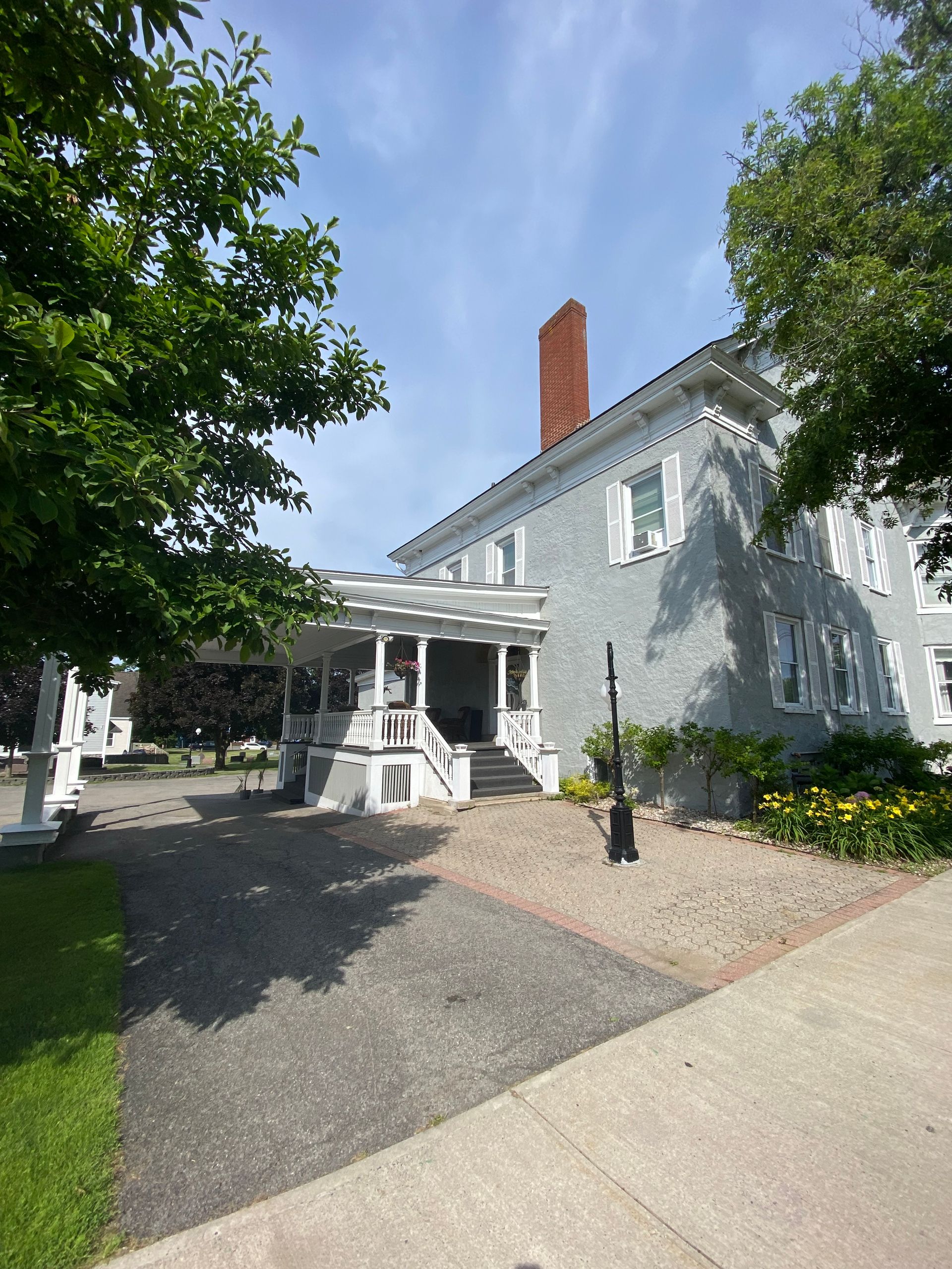 A light gray, historic two-story building with a red brick chimney, a white covered porch, and landscaped gardens.