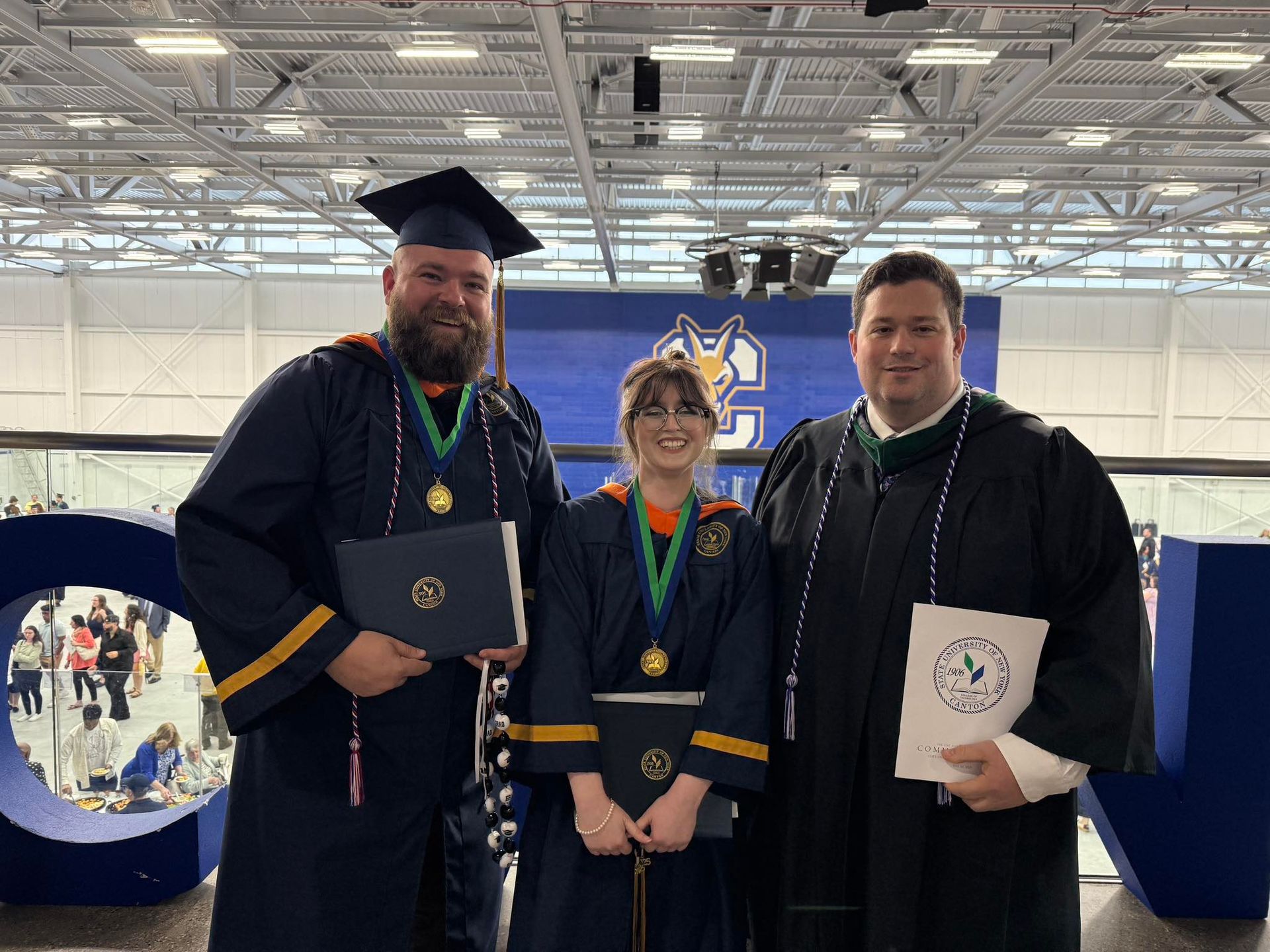 Three people in academic regalia holding diplomas and awards stand in front of a blue college banner at a graduation.