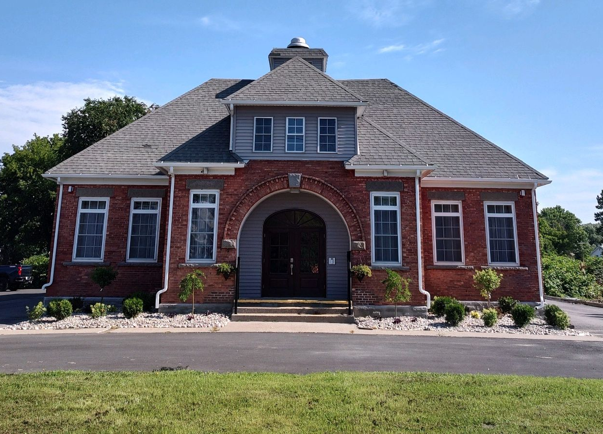 A brick building with a large arched entrance, gray shingles, and a central chimney set against a blue sky.
