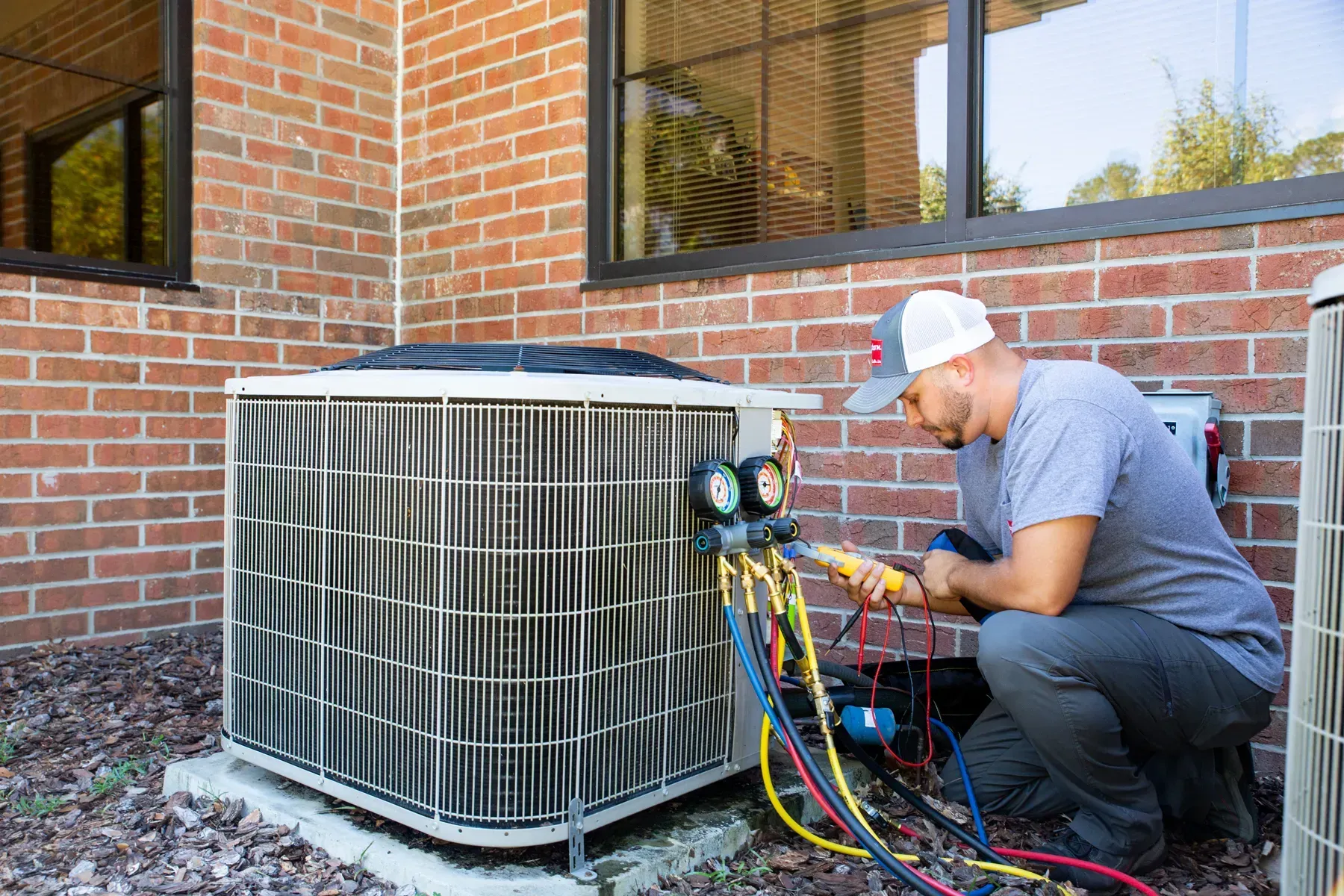 A person is servicing an air conditioning unit. Outdoors, near brick wall, using gauges and hoses.