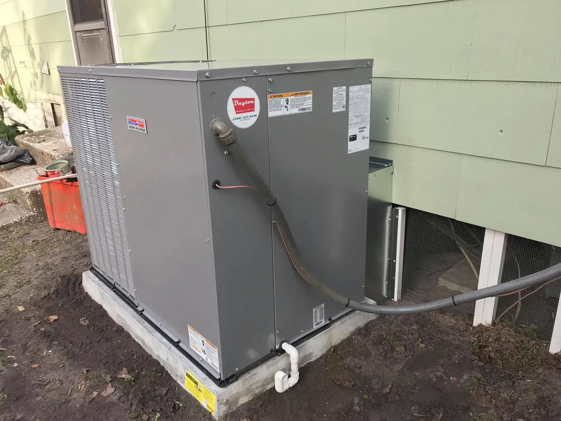 A gray air conditioning unit installed outside a building on a concrete pad with a conduit and PVC pipe.