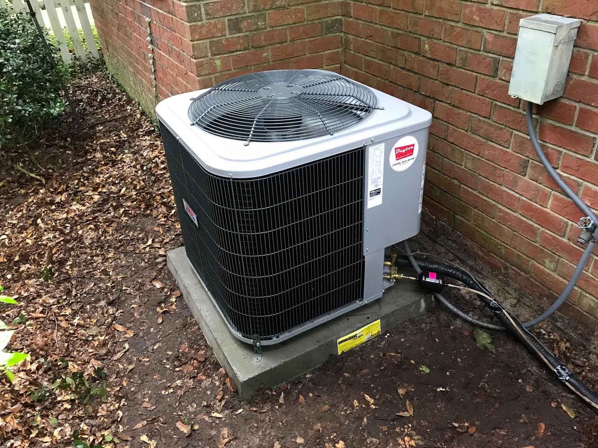An air conditioning unit on a concrete pad near a brick wall and a fence.