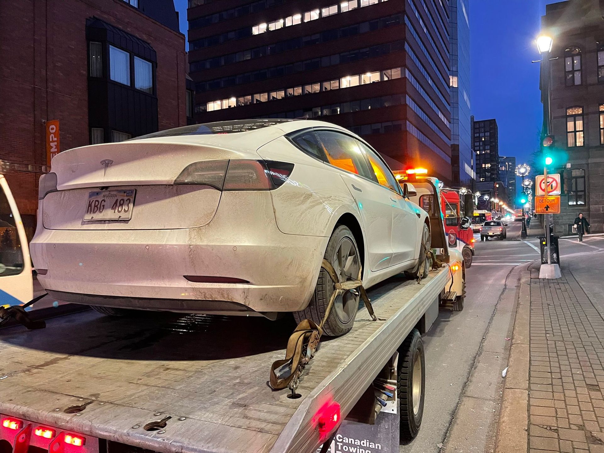 White sedan loaded on a tow truck at night on a city street, with buildings and traffic lights nearby