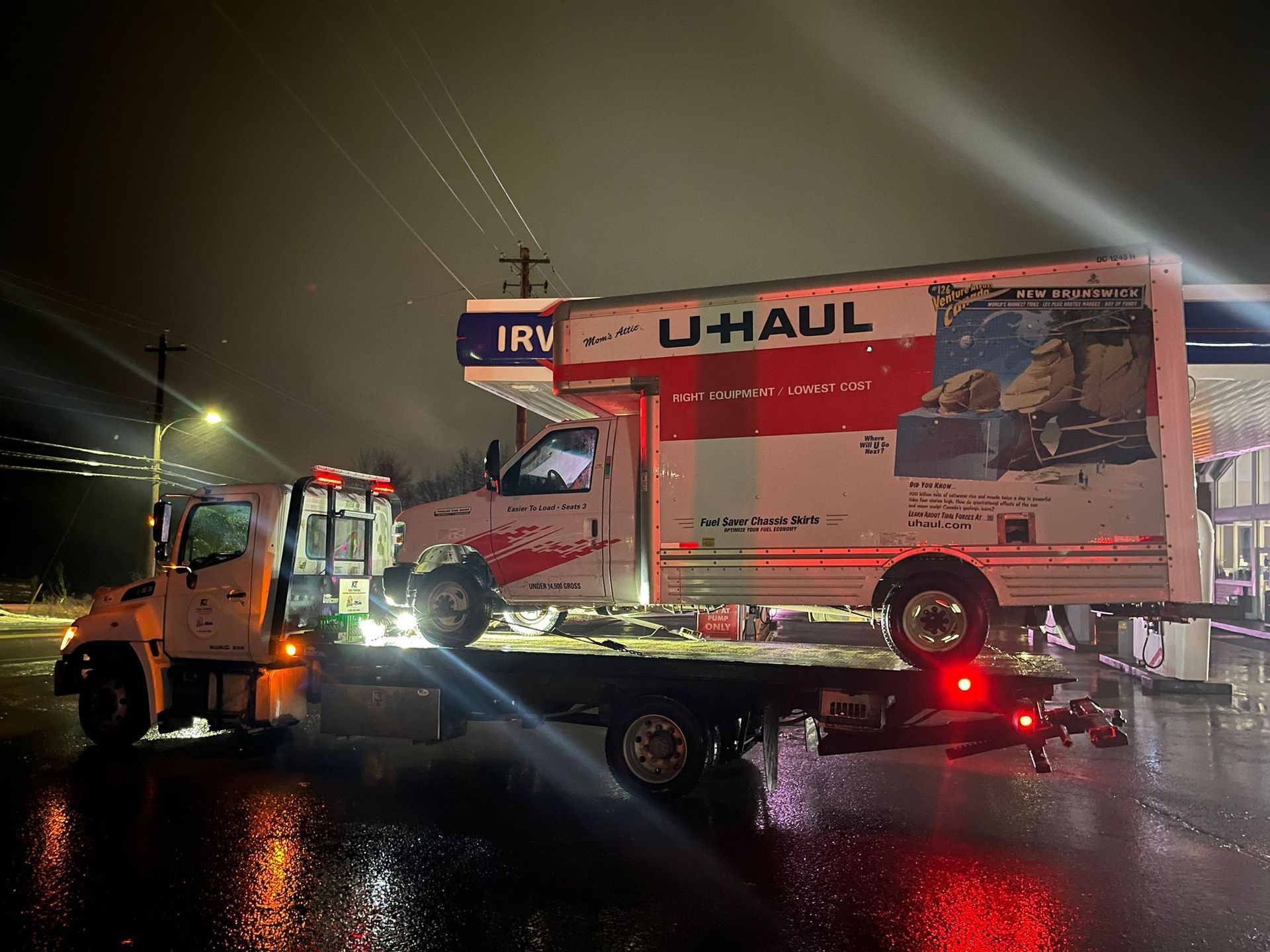 Tow truck hauling a U-Haul box truck on a wet road at night with flashing lights