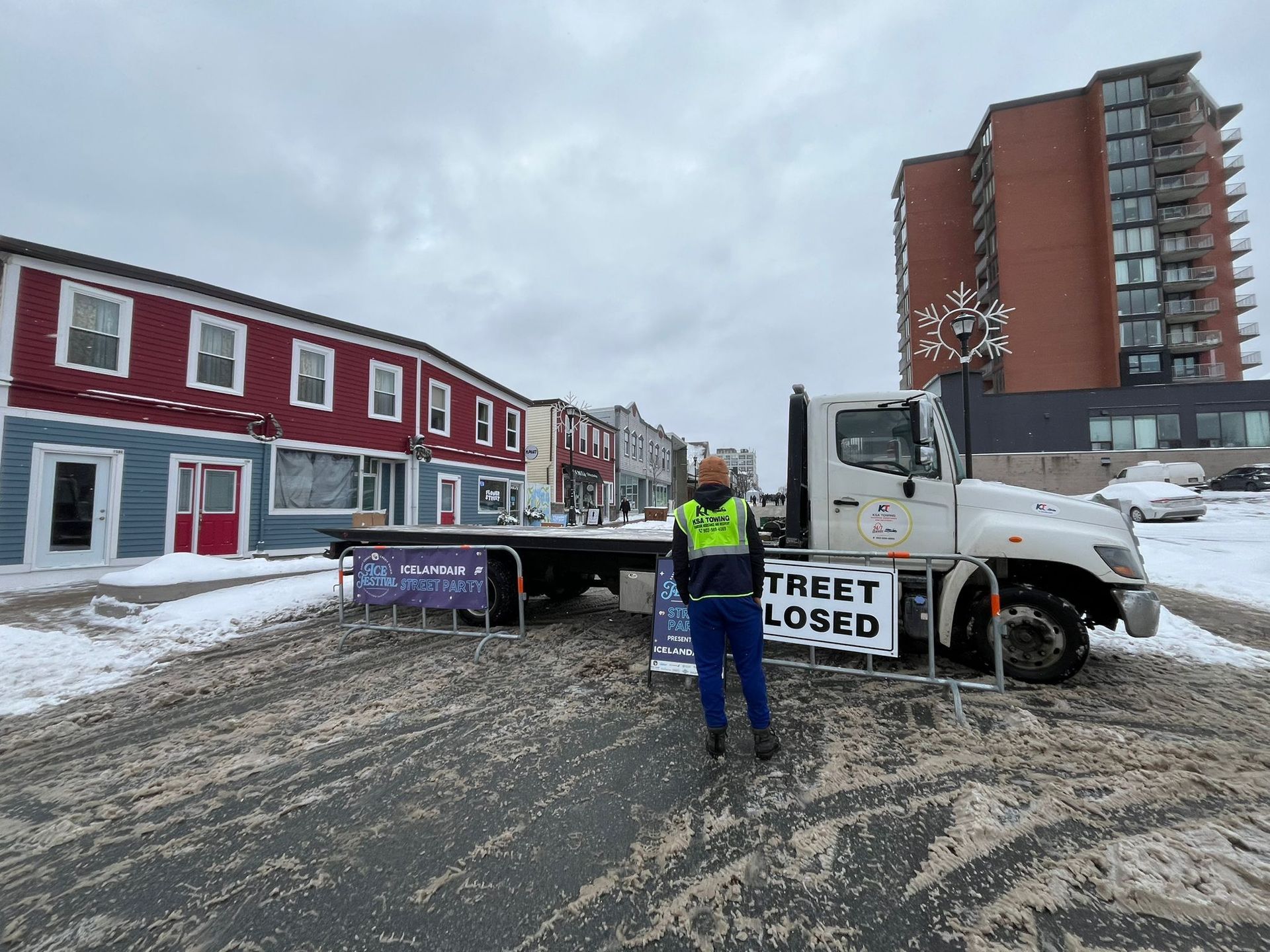 Snowy street with road closed sign, worker in safety vest, truck, and damaged buildings.