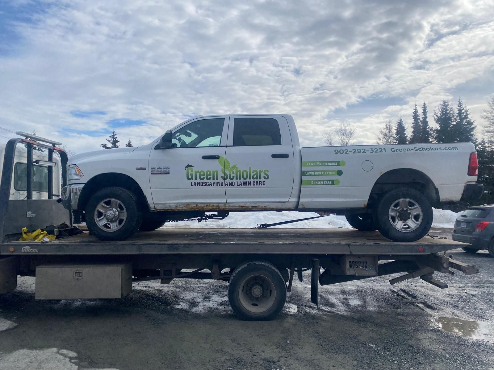 White pickup truck with company logo on a flatbed trailer in a snowy lot under cloudy sky