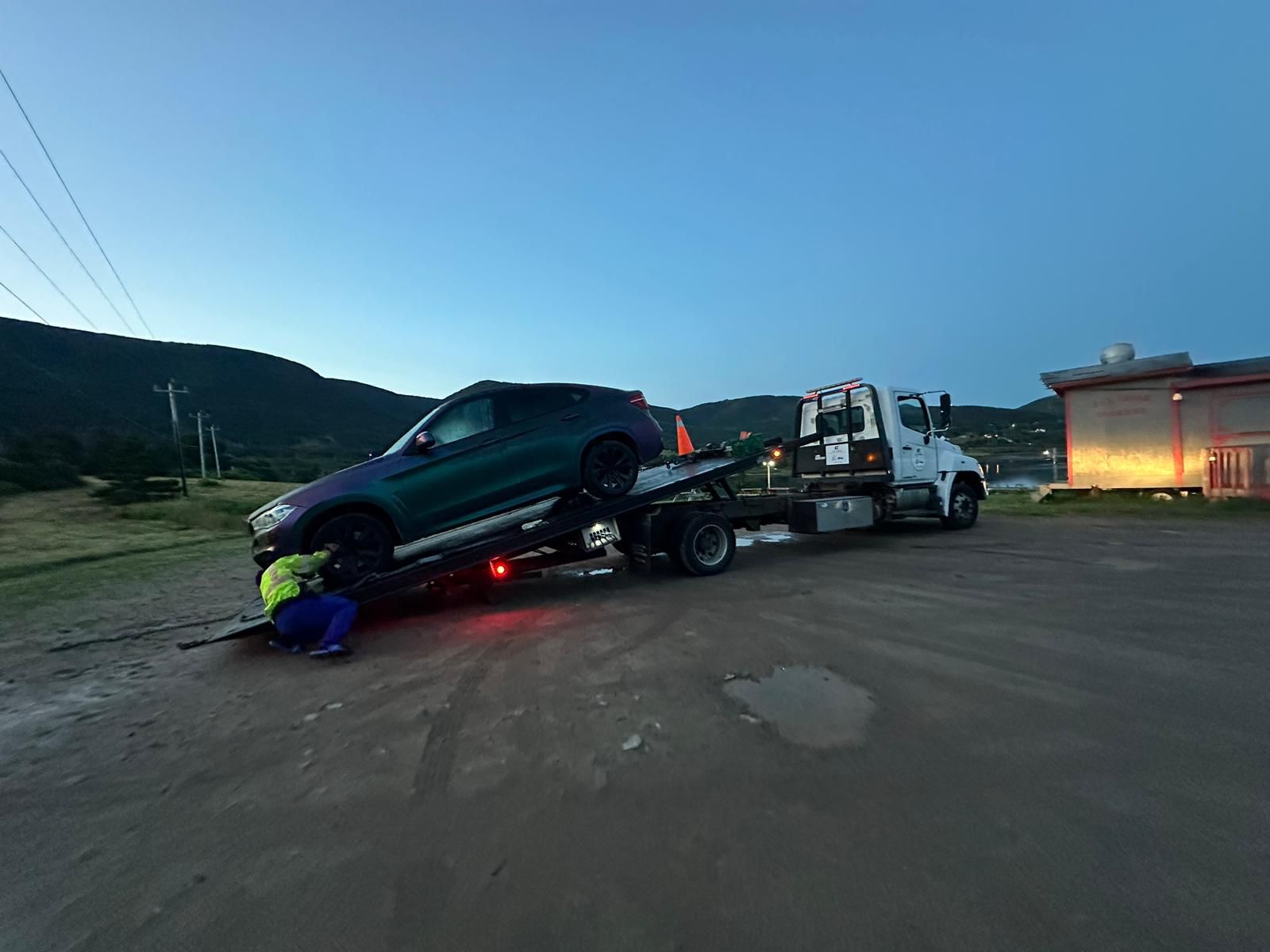 Tow truck loading a dark SUV at dusk in a roadside lot, with red lights glowing under the truck.