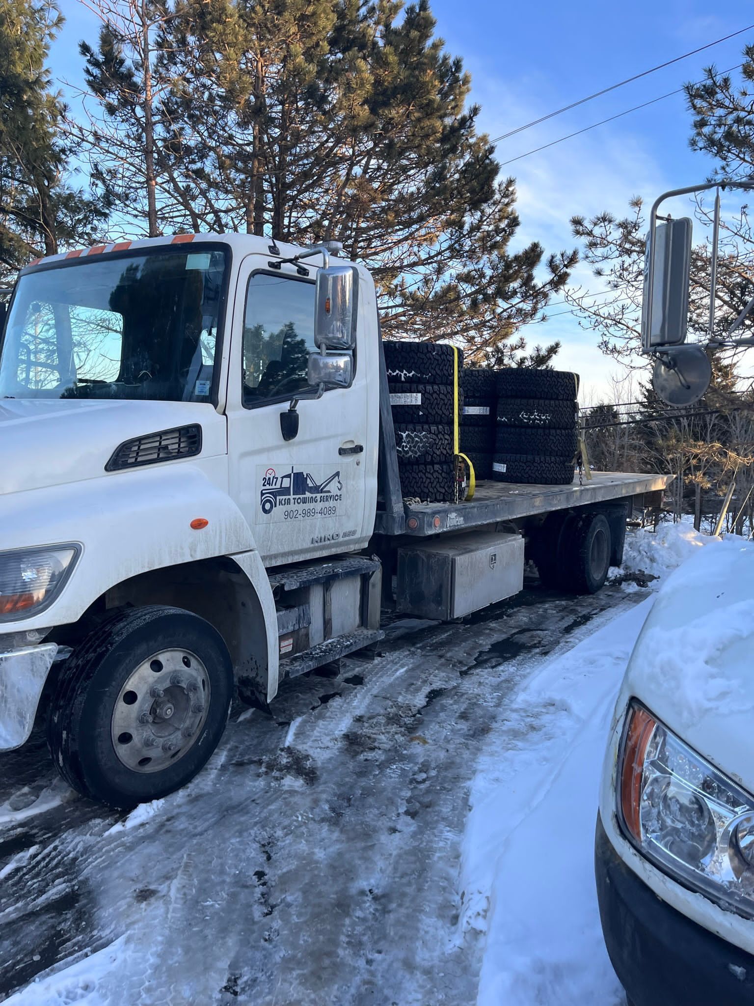 White flatbed truck carrying black crates on a snowy road beside parked cars and trees