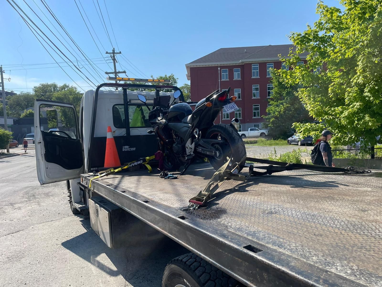 Tow truck carrying a motorcycle on a flatbed, with a traffic cone and red brick building in the background