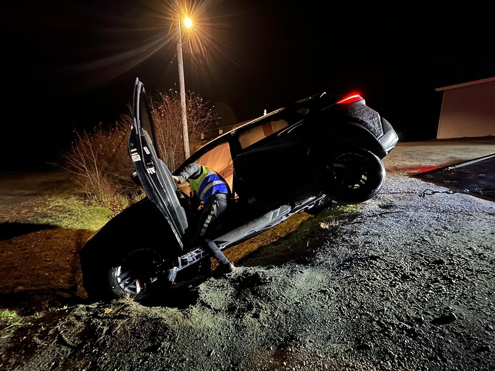 Overturned black car in a ditch at night, with a deployed airbag and a firework-like burst nearby.