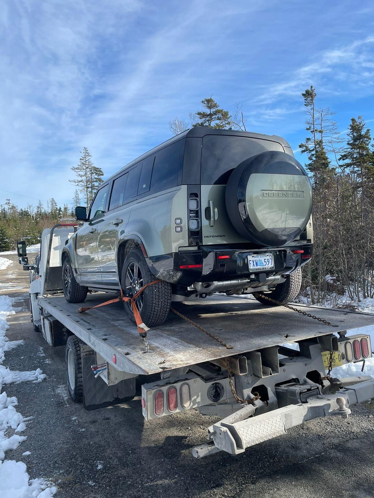 Gray SUV on a flatbed tow truck with rear wheel strapped down on a snowy roadside.