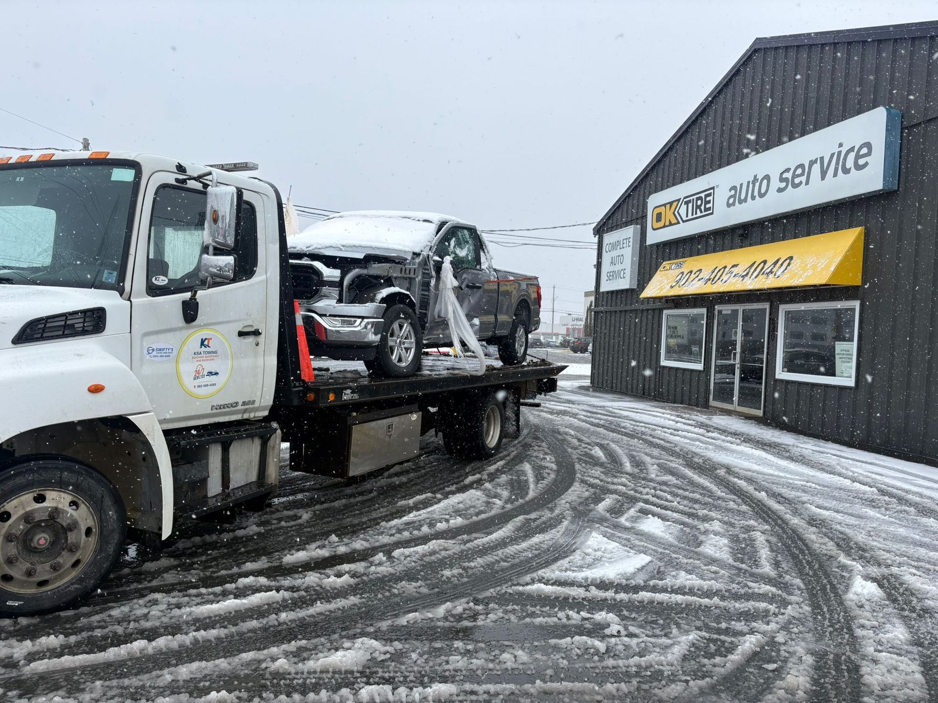 Tow truck carrying a damaged SUV outside an auto service shop on a snowy street