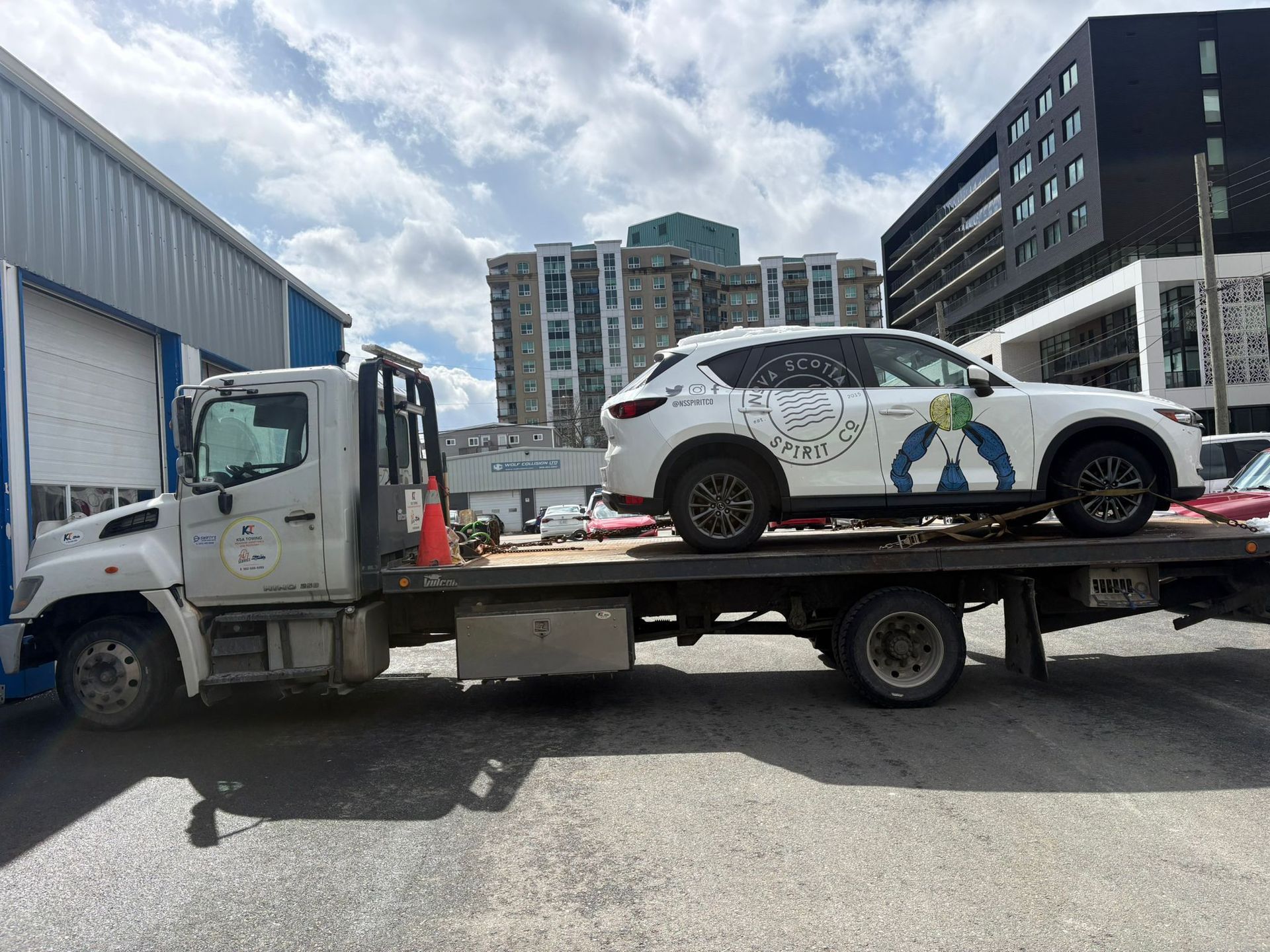 Tow truck carrying a white car with colorful graphics in a parking lot between buildings