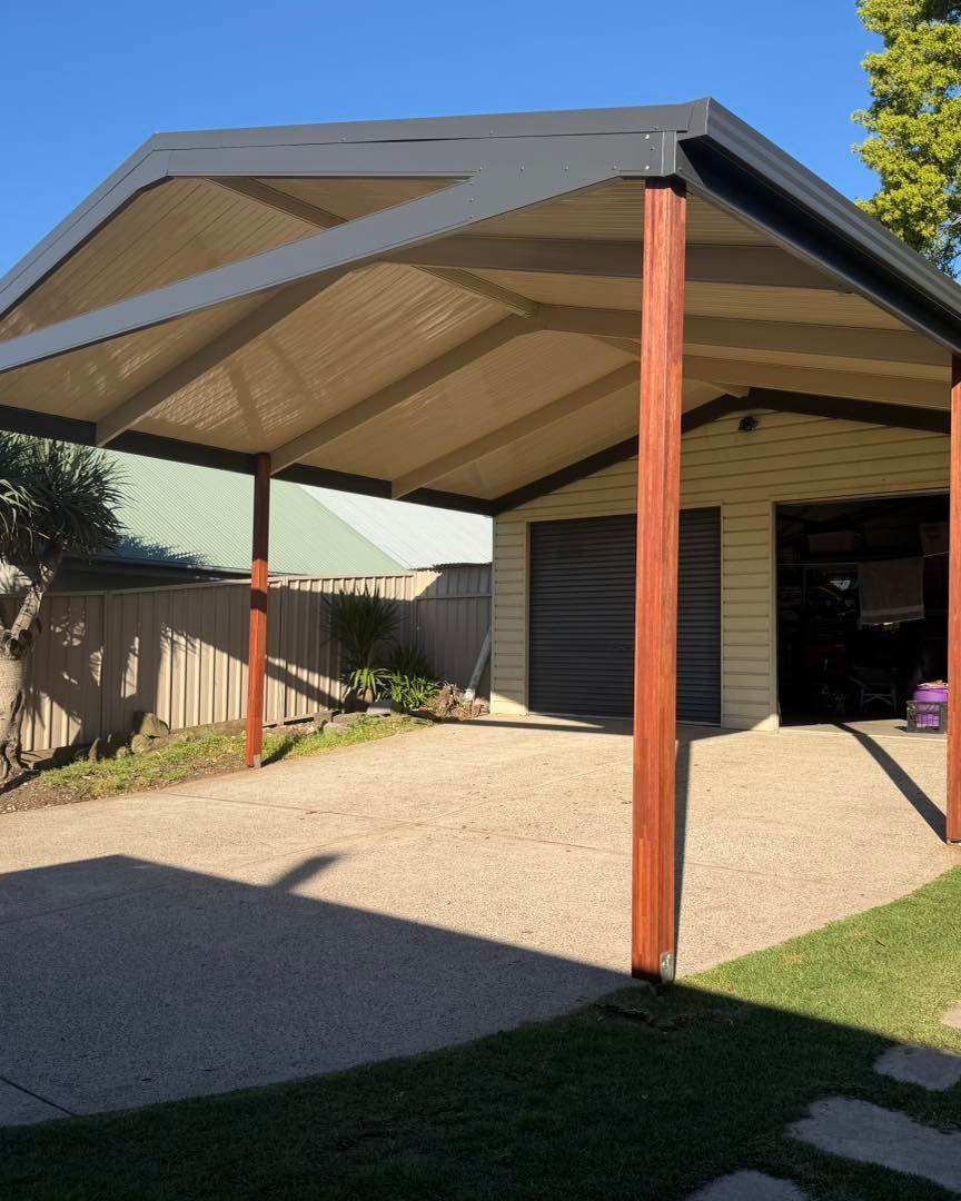 Freestanding carport with beige roof and red posts beside a house, over a gravel driveway