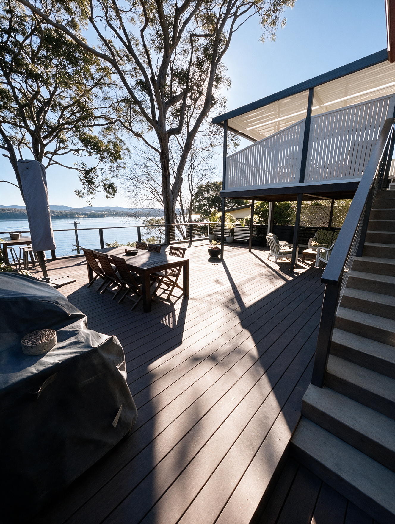 Wooden lakeside deck with stairs, picnic tables, and trees in bright sunlight.