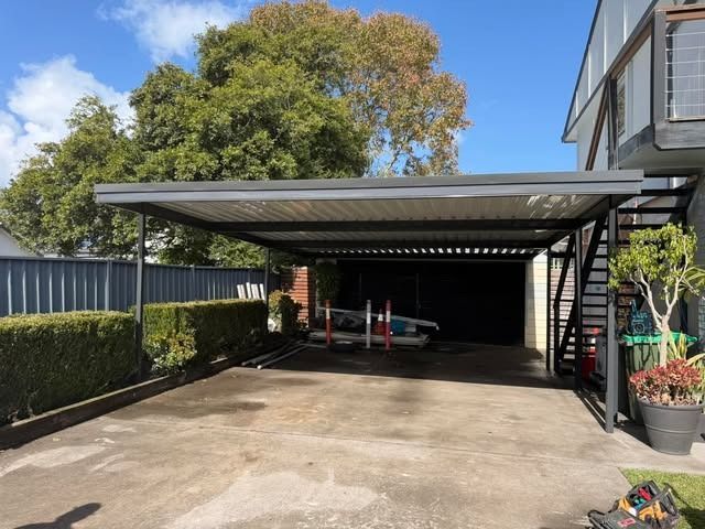 Carport driveway beside a house, with a metal roof, hedge fence, and potted plants.