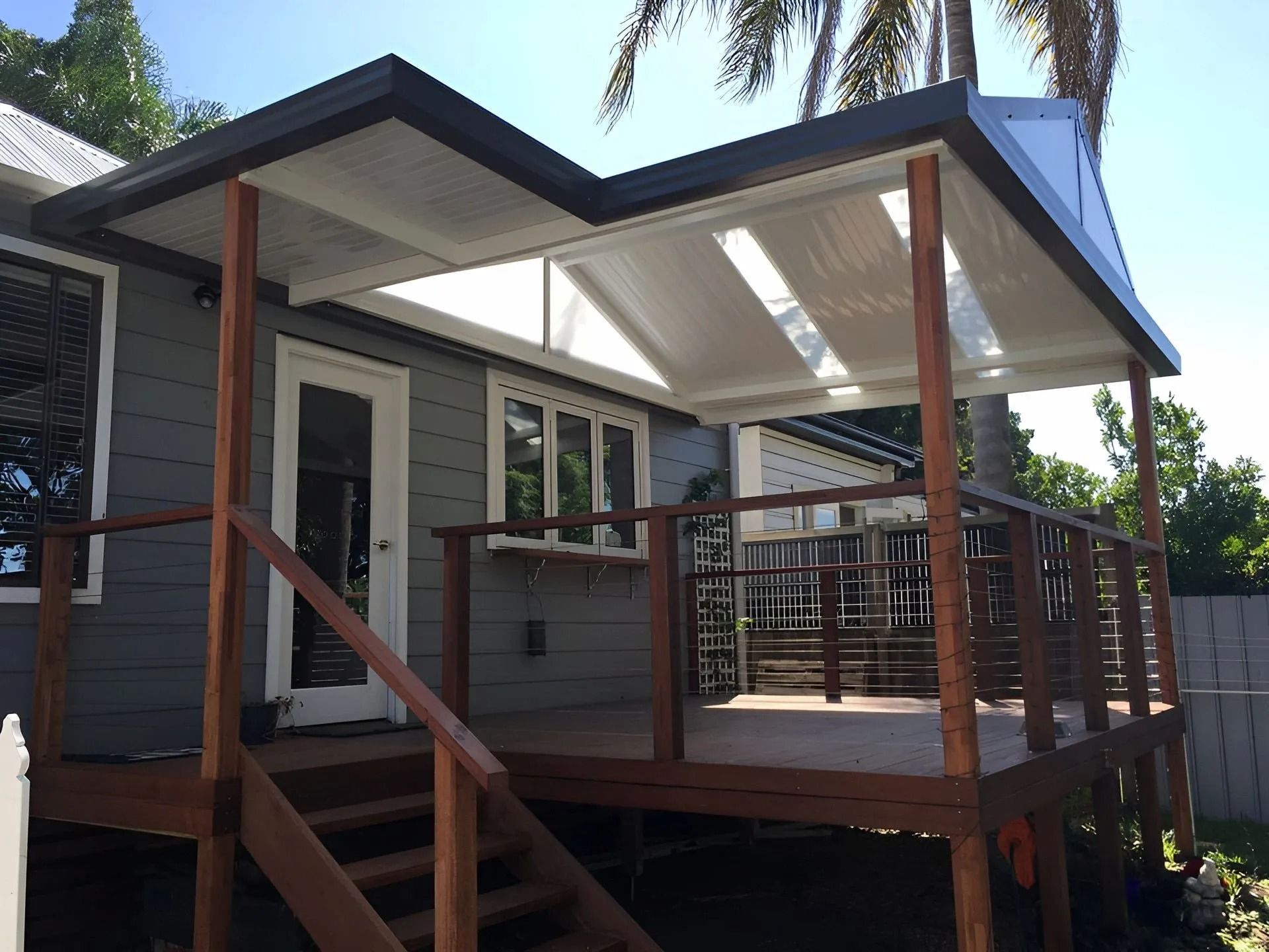 Small wooden house deck with railing and stairs, covered by a white roof in a sunny yard
