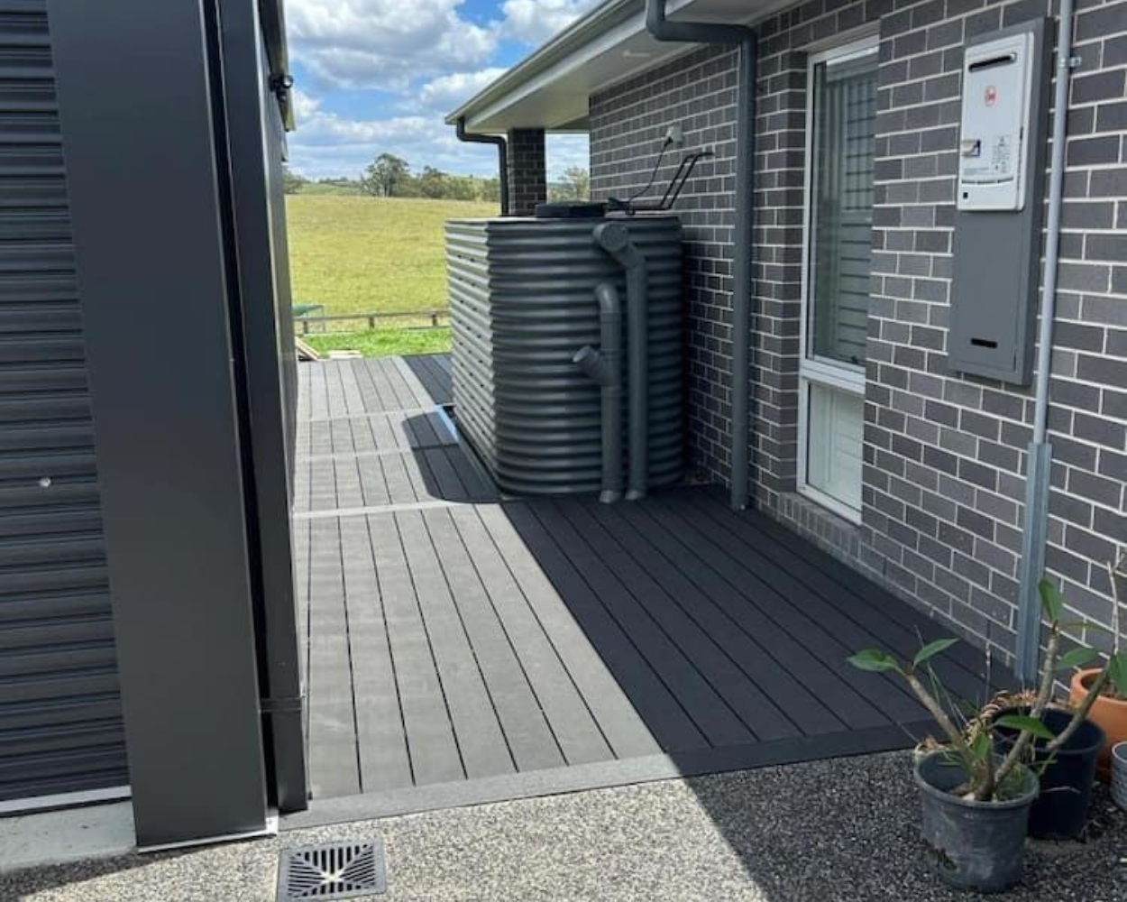 Exterior side walkway beside a brick house, with stacked black water tanks and potted plants.