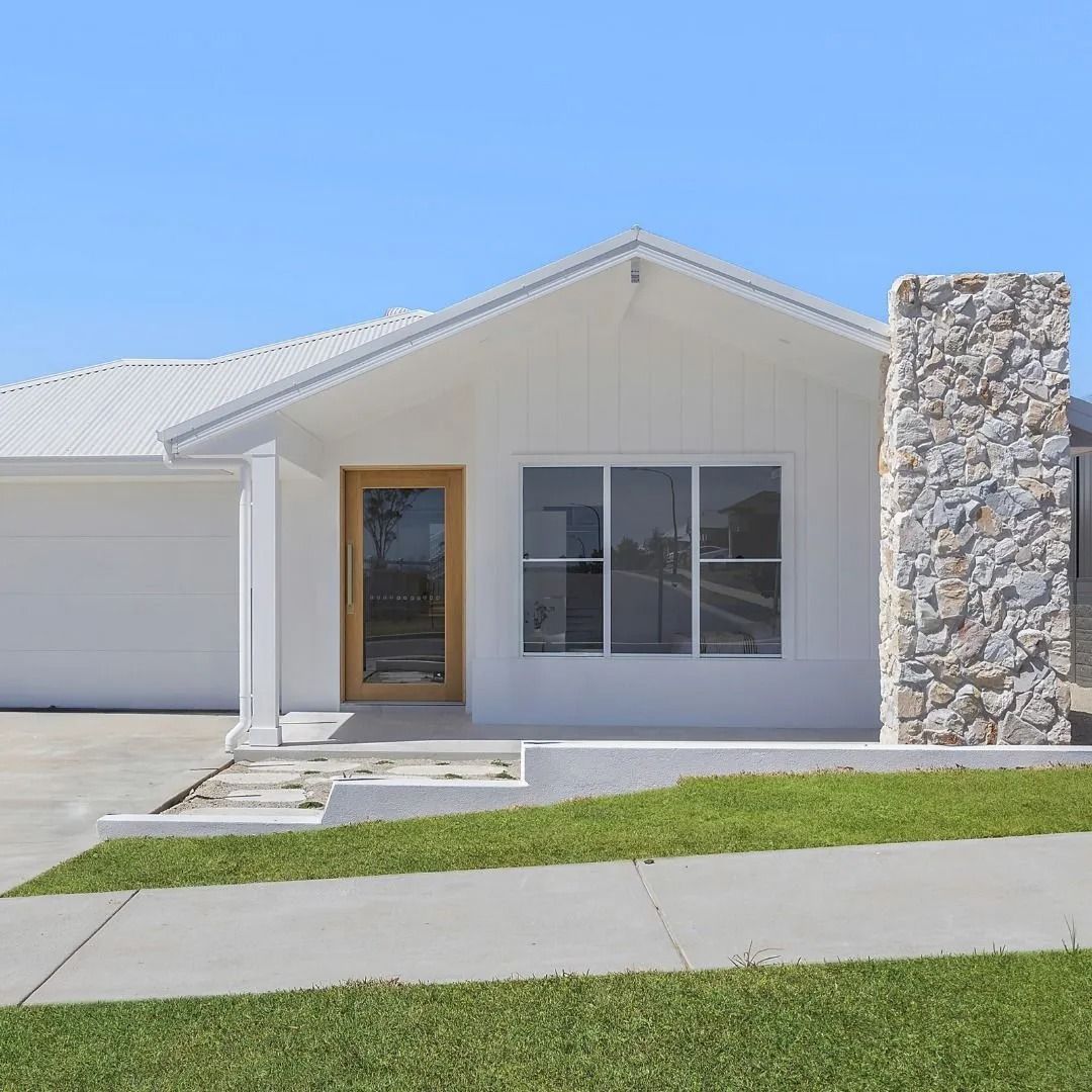 White house front with stone column, garage, walkway, and green lawn under a blue sky