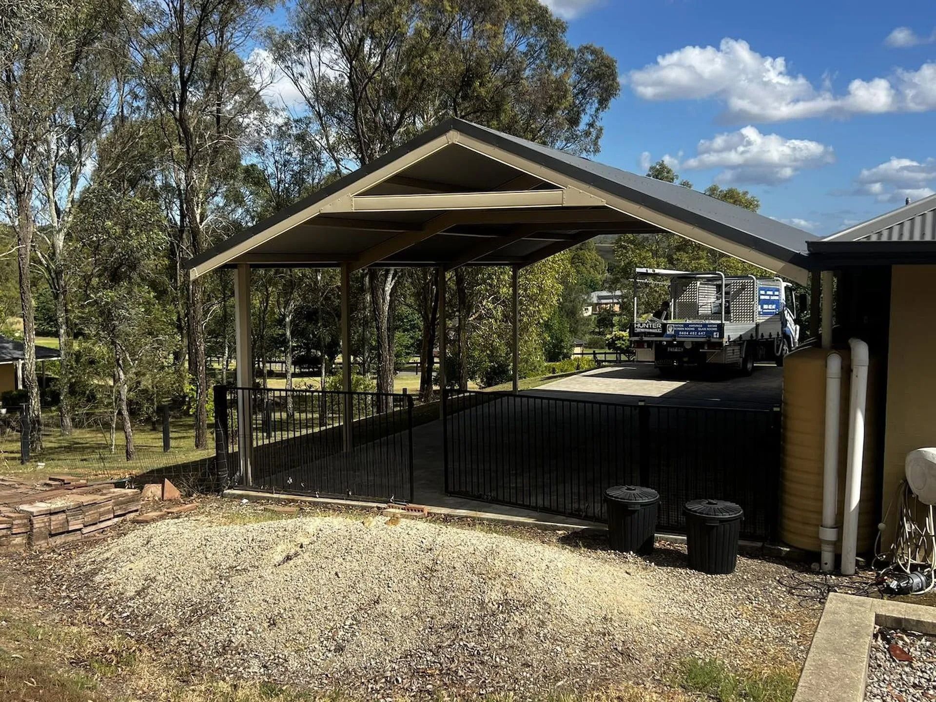 Covered driveway beside a house with a truck, black fence, and gravel pile under a blue sky