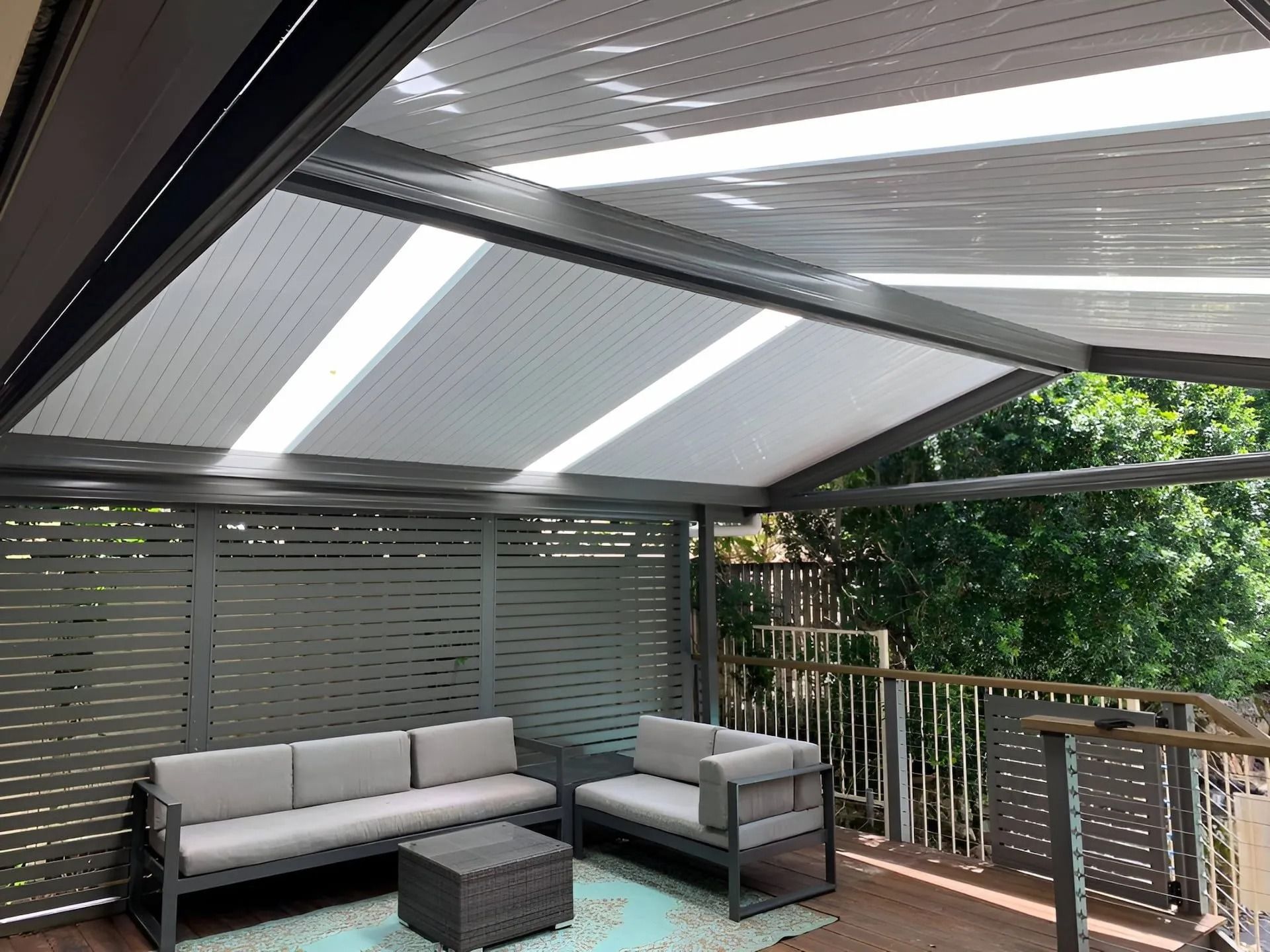 Covered patio with gray outdoor sofa, matching chairs, and a dark coffee table under slatted roof panels.
