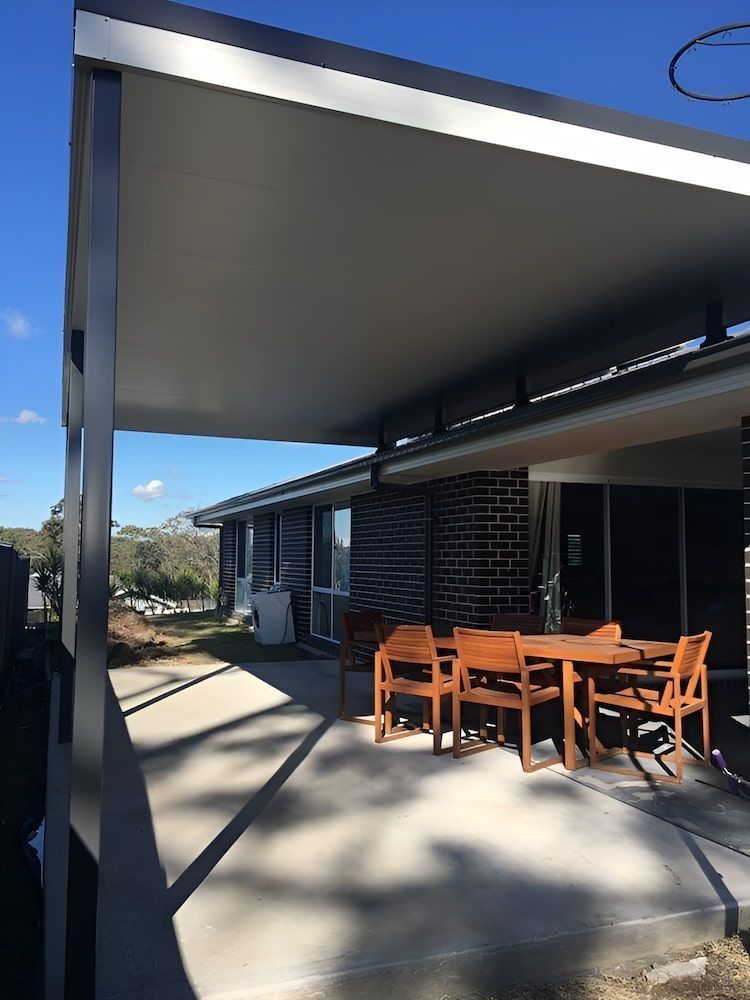 Covered patio with wooden dining table and chairs beside a modern house, lit by sunlight.