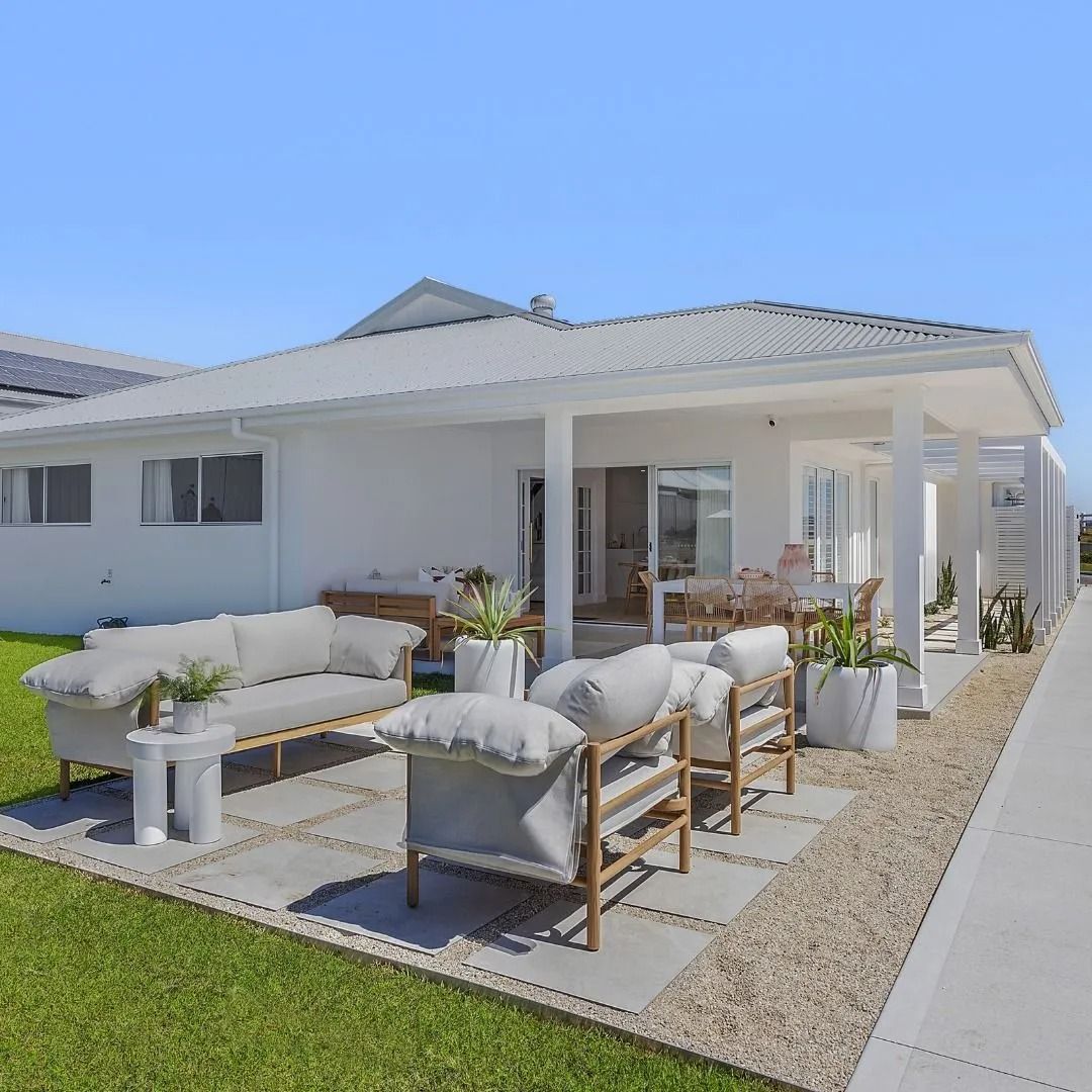 Modern white patio with outdoor sofa, chairs, and dining table beside a white house under blue sky