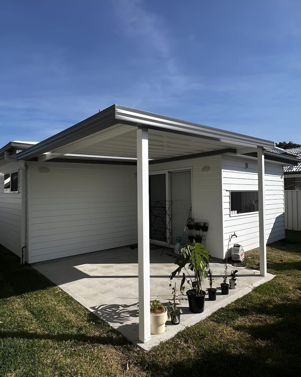 Small white carport beside a white house, with potted plants on the concrete patio under a metal roof.