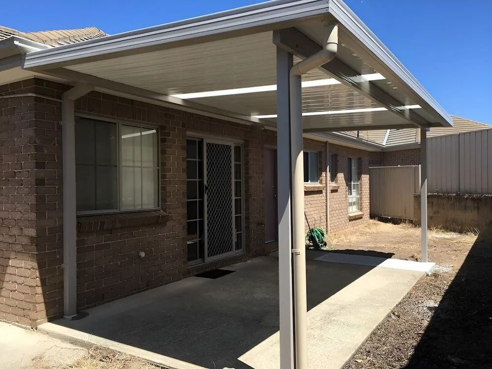 Covered brick patio with metal posts beside a house, shaded concrete walkway, and clear blue sky.