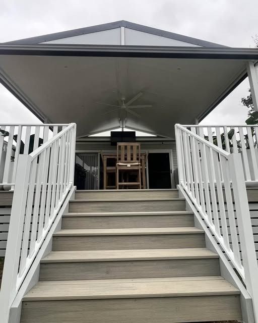 Covered front porch with white stairs, railing, ceiling fan, and seating beneath a peaked roof.