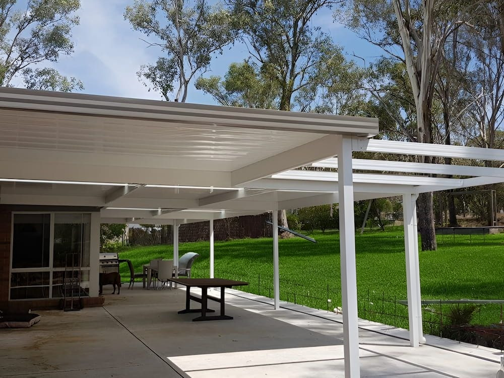 Covered outdoor walkway with white posts, picnic tables, and a green lawn beside trees