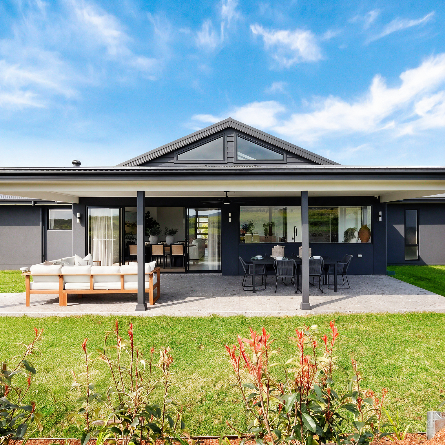 Modern single-story house with a covered patio, outdoor seating, and a green lawn under a blue sky