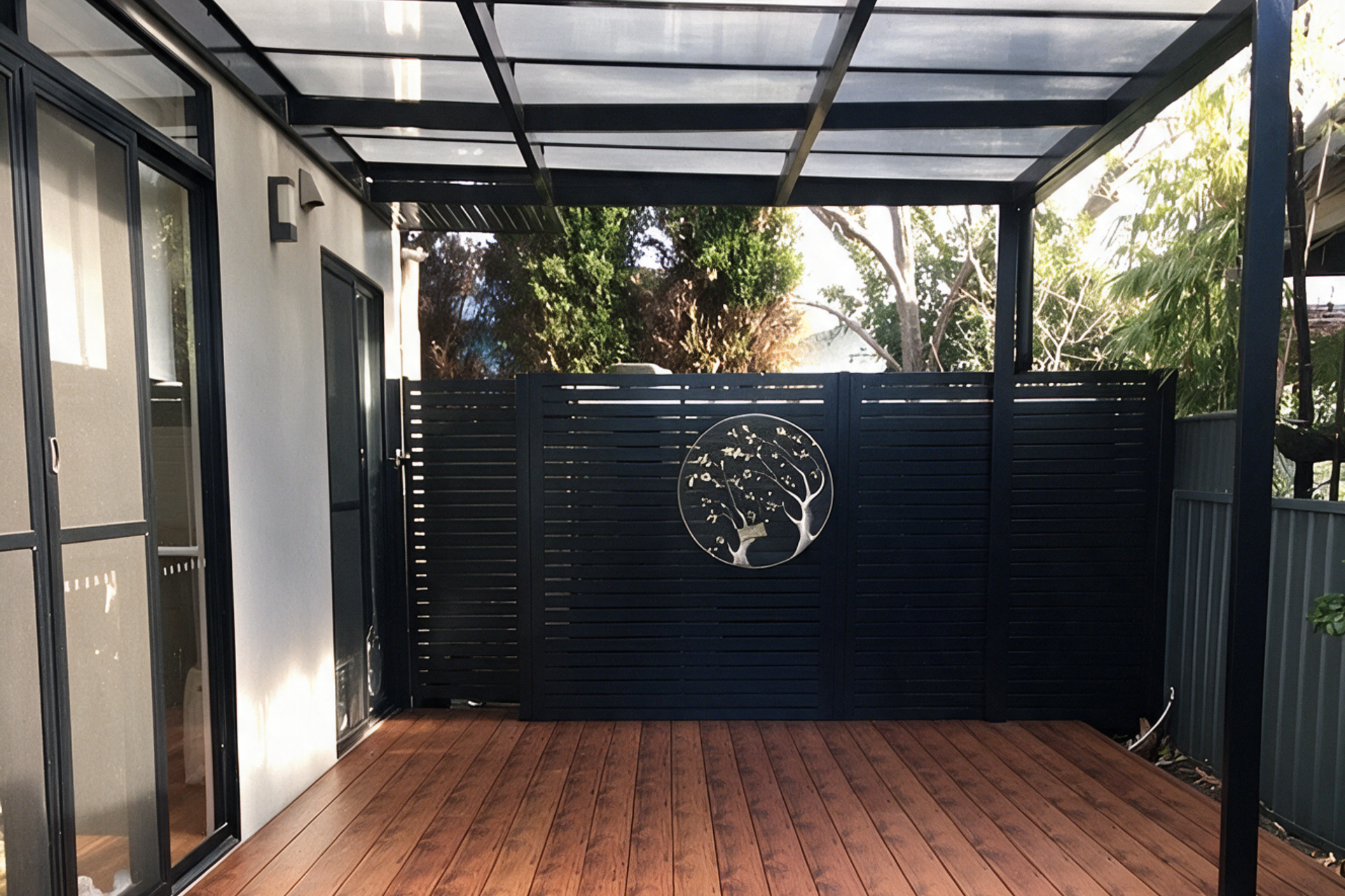 Covered patio with wooden deck, black metal posts, and a dark privacy screen with a circular emblem.